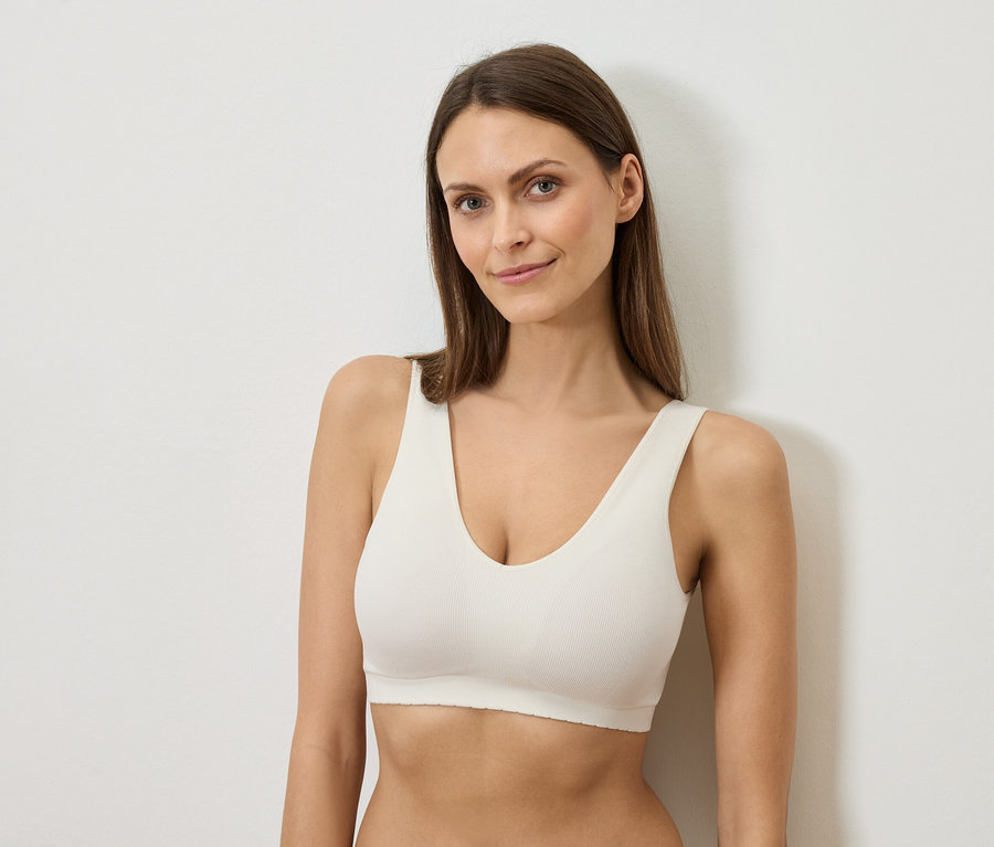 Žena s dlouhými hnědými vlasy v bílé podprsence se opírá o bílou zeď.