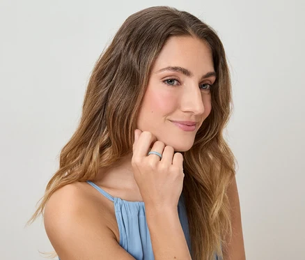 Žena s vlnitými vlasy a v modrém topu se dotýká prstu s prstenem na světlém pozadí.