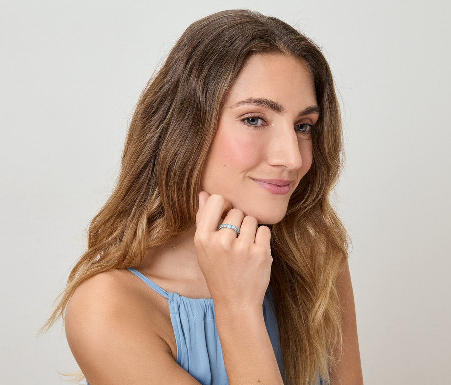 Žena s vlnitými vlasy a v modrém topu se dotýká prstu s prstenem na světlém pozadí.