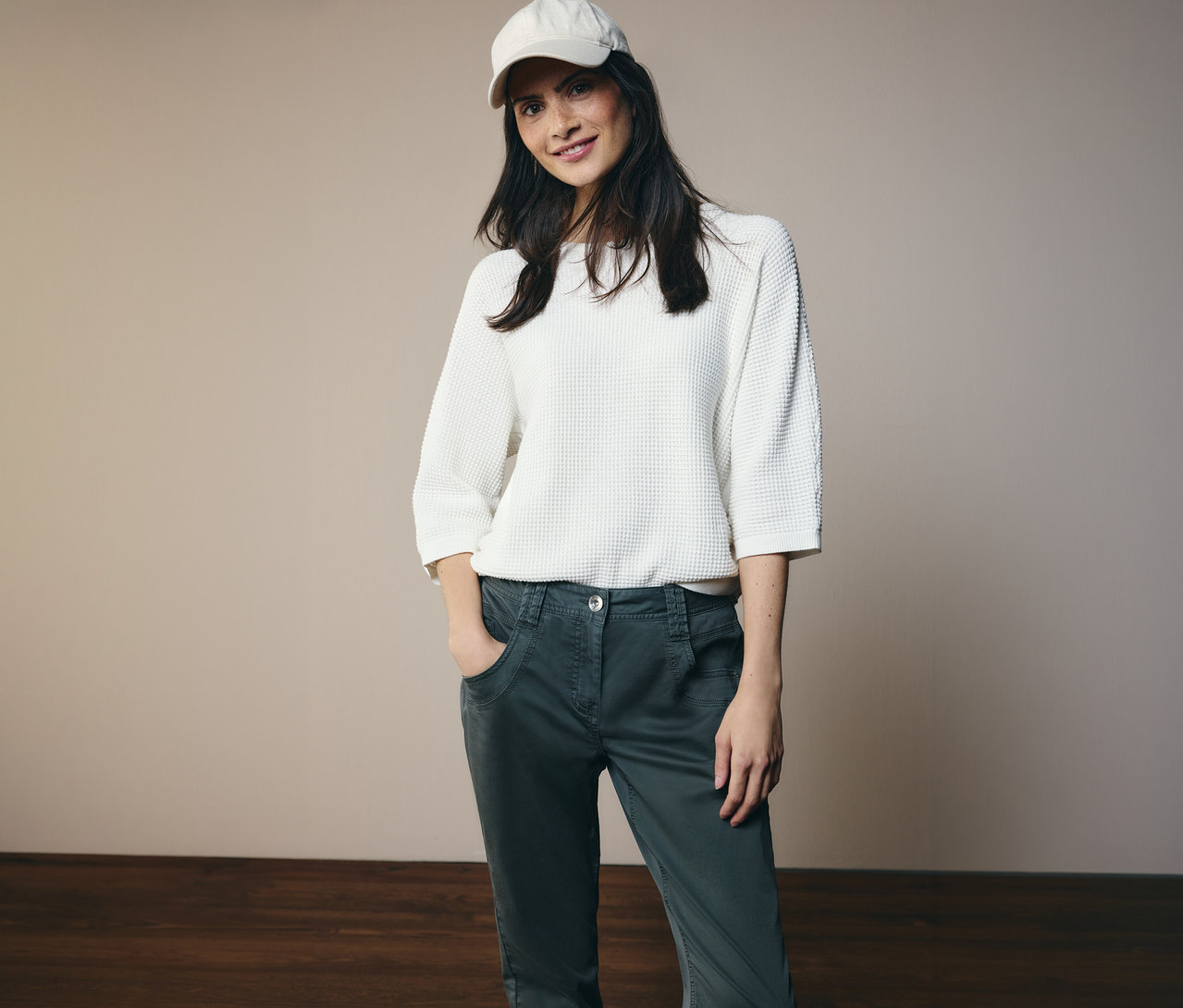 Žena v bílém topu a čepici pózuje v kalhotách CECIL »Scarlett« s rukama v kapsách.