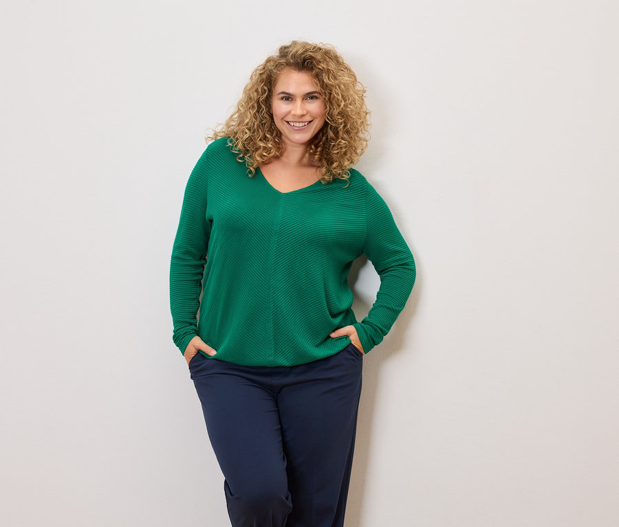 Žena s kudrnatými vlasy má na sobě zelený svetr a tmavě modré kalhoty, ruce v kapsách.