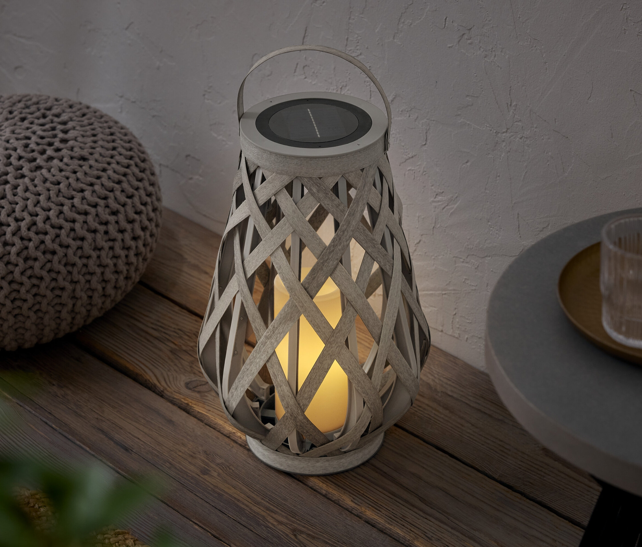 Pletená lucerna se solárním panelem stojí vedle pufu na dřevěné podlaze.