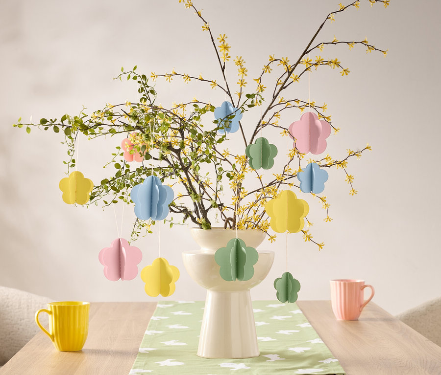 Květinové ozdoby visí na větvi ve váze na stole, vedle žlutého a růžového hrnku.