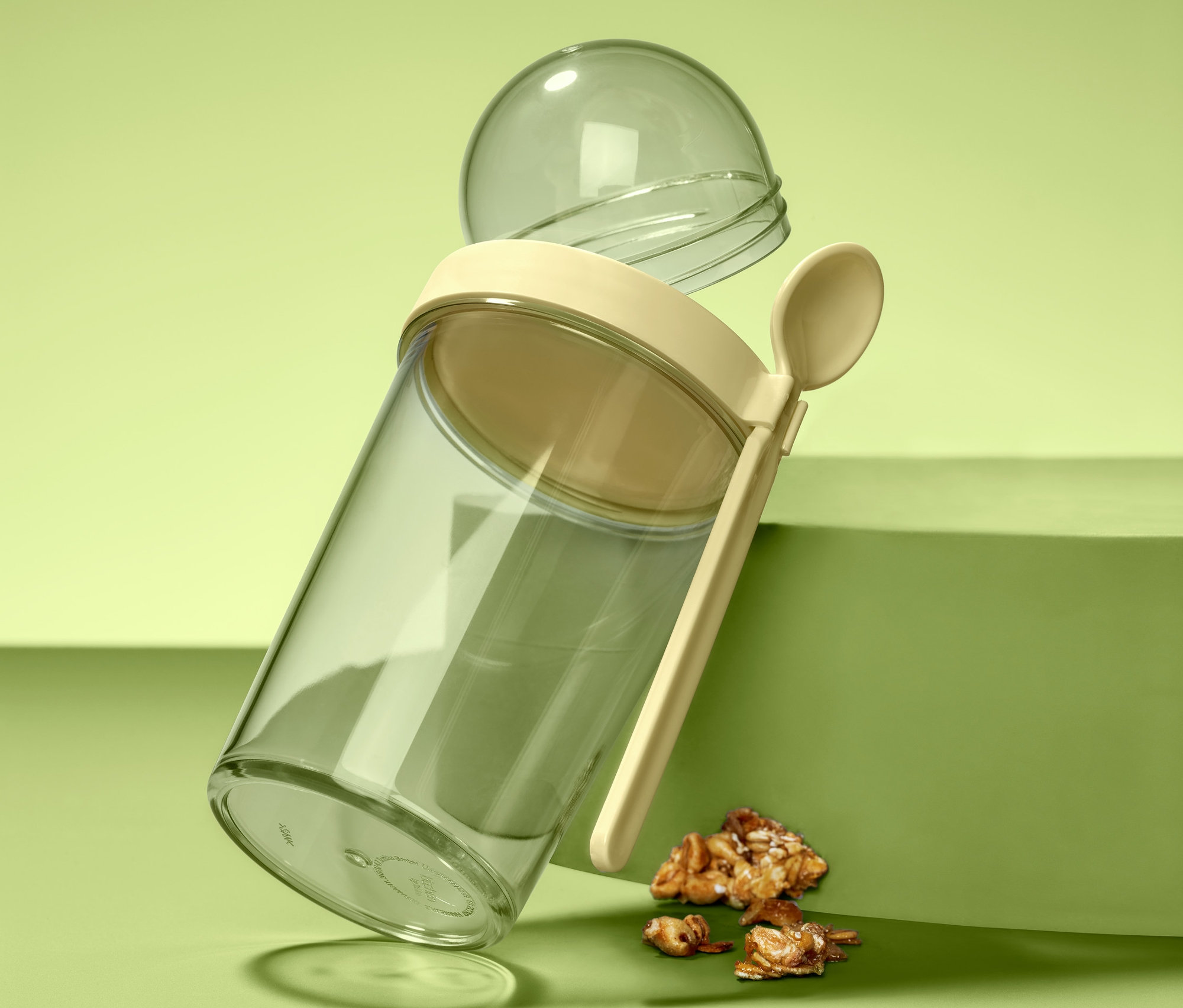 Průhledný cestovní kelímek na jogurt s béžovým víčkem, lžící a průhlednou kulovou nádobou stojí nakloněný na zeleném povrchu vedle kousků müsli.