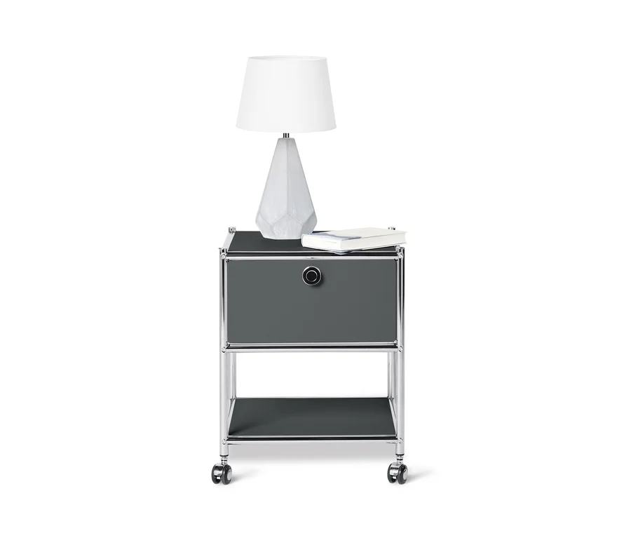 Šedý kovový odkládací stolek CN3 se zásuvkou, na kterém stojí bílá lampa a kniha.