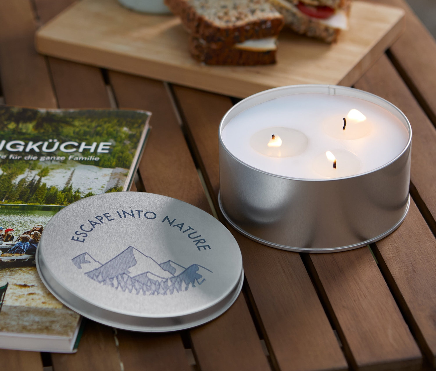 Na dřevěném stole leží svíčka citronella, kniha Campingküche, hrnek a sendviče.