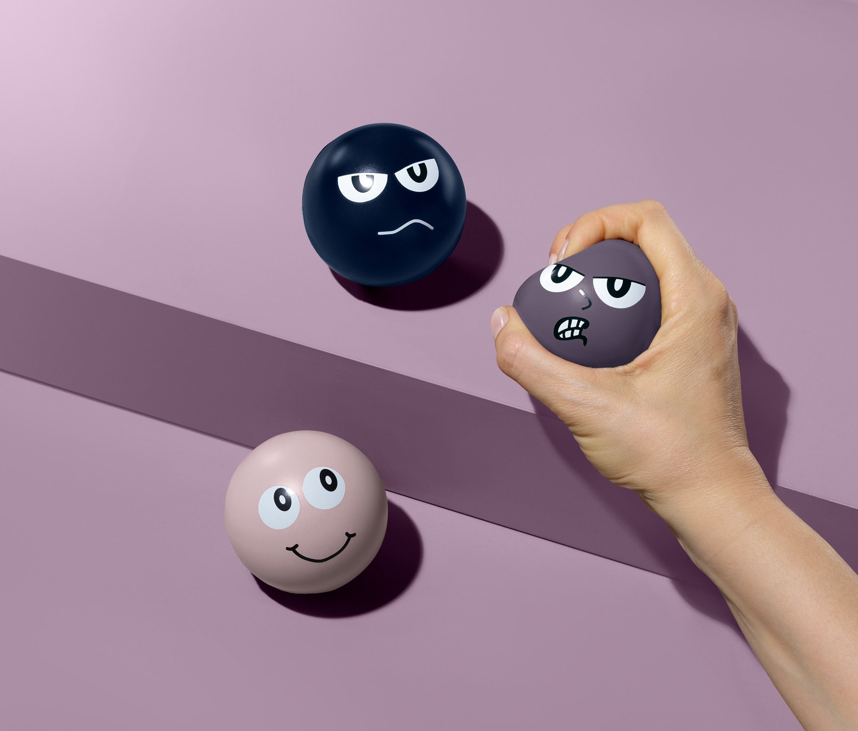 Ruka mačká fialový antistresový míček se zamračeným obličejem. Na fialovém pozadí leží další dva antistresové míčky.