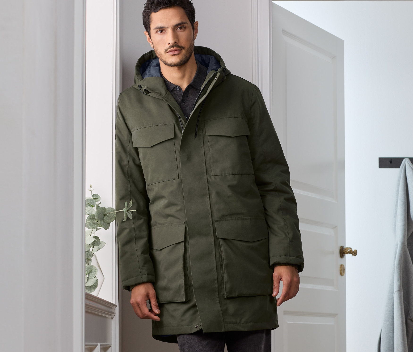 Muž opřený o zeď má na sobě olivově zelenou parku.