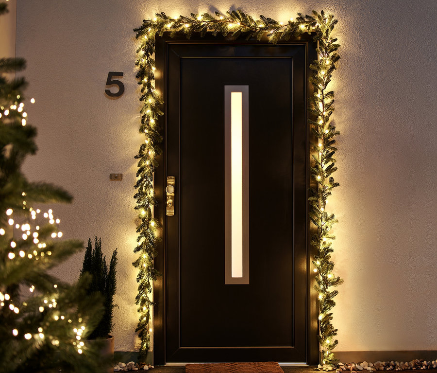 Vchodové dveře orámované jedlovou girlandou s LED světly, vlevo vánoční stromek s LED světly.