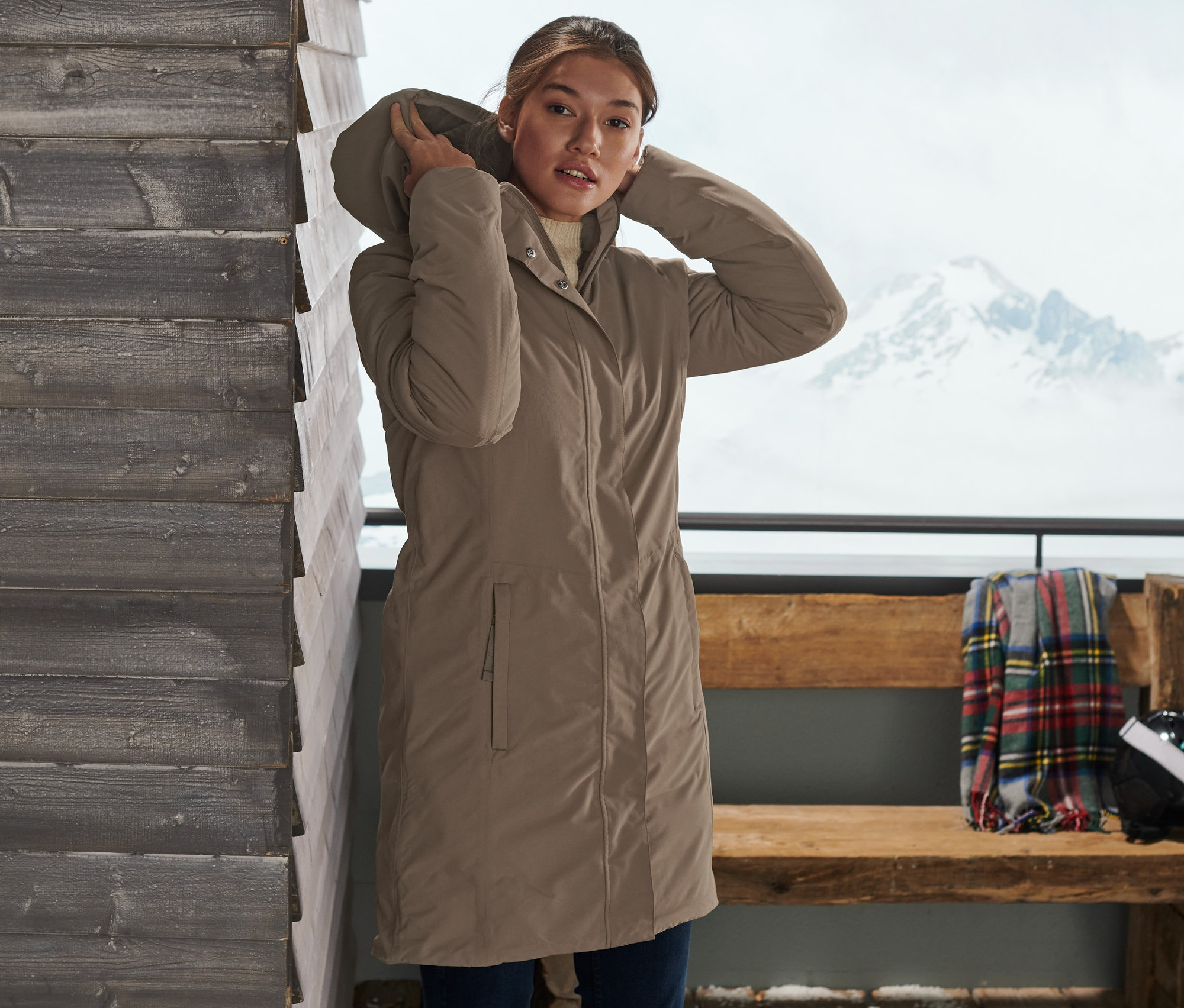 Žena si upravuje kapuci šedobéžového termokabátu. V pozadí jsou lyže a zasněžená krajina.