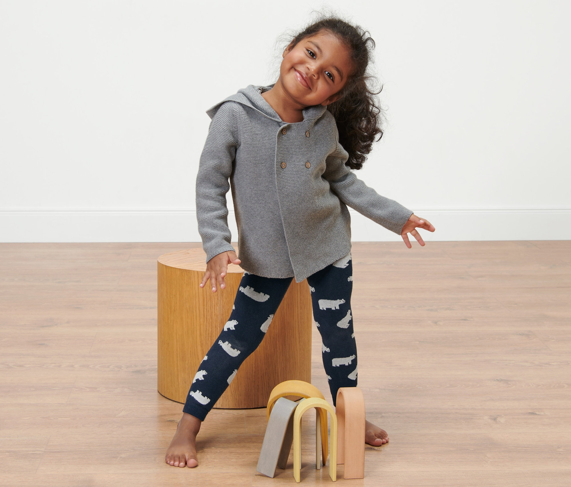 Usmívající se holčička stojí a má na sobě dětské termolegíny, 2 ks s potiskem ledních medvědů a šedý kabátek.