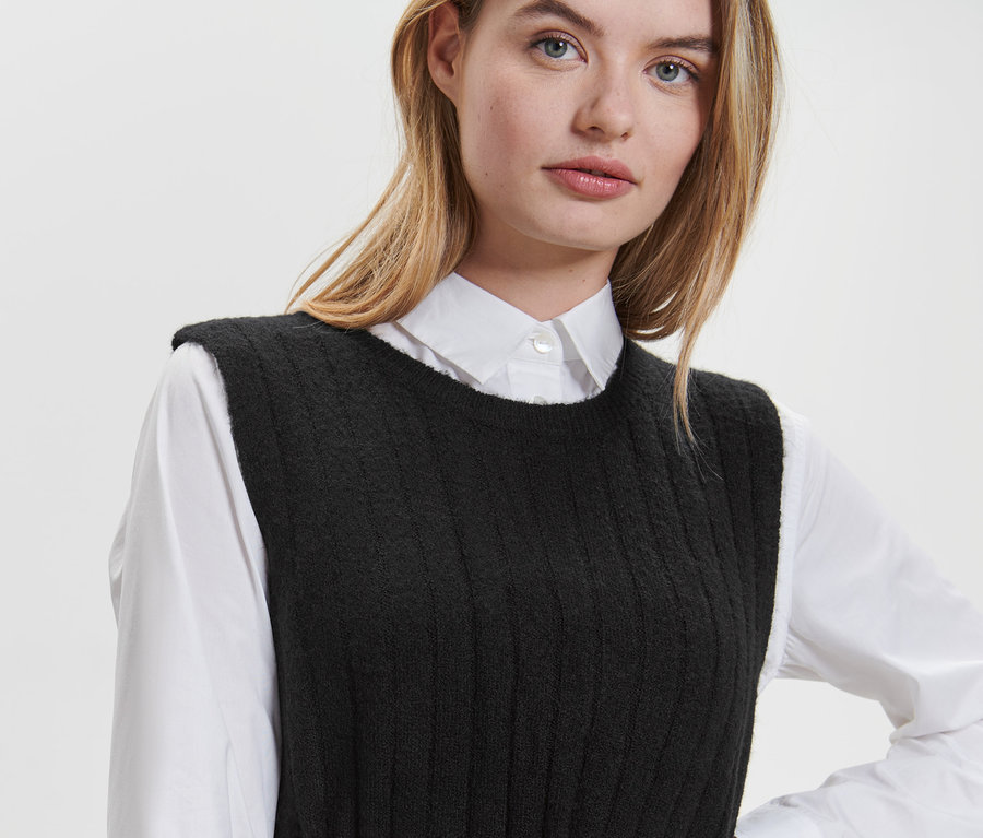 Blondýna má na sobě černé žebrované pletené šaty a bílou košili.