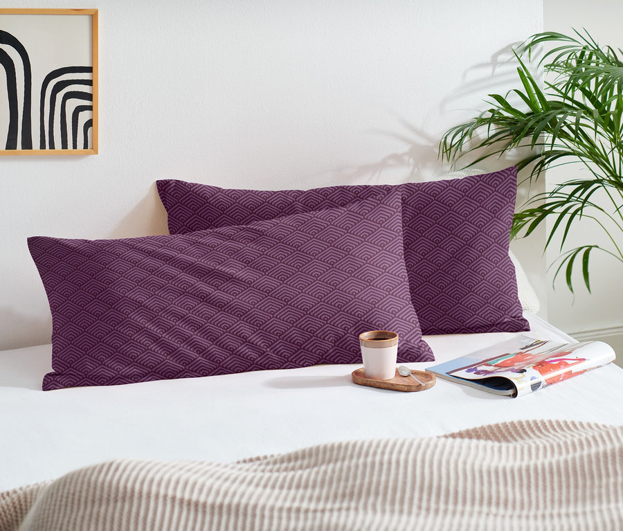 Dva biberové povlaky na polštáře s fialovým vzorem na posteli s šálkem a časopisem.