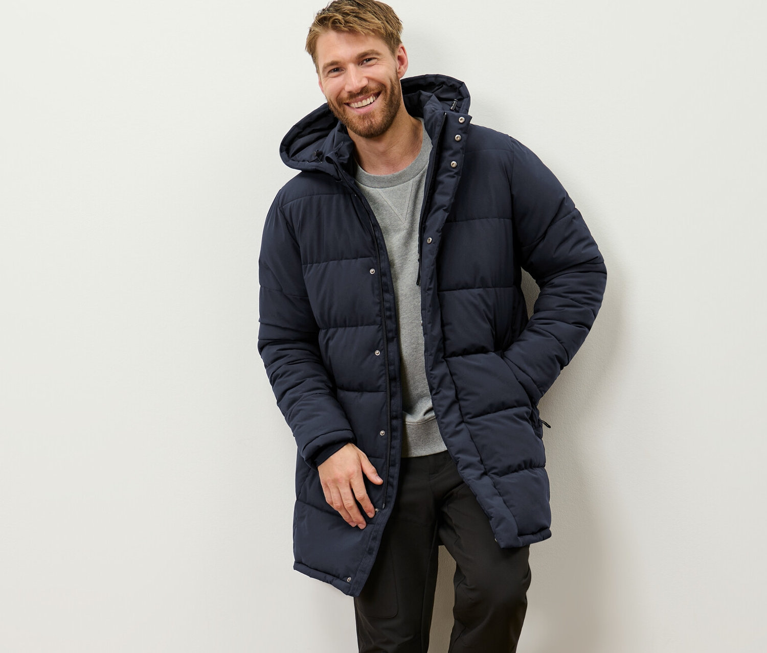 Muž se usmívá a opírá se o stěnu v prošívané parke s kapucí.