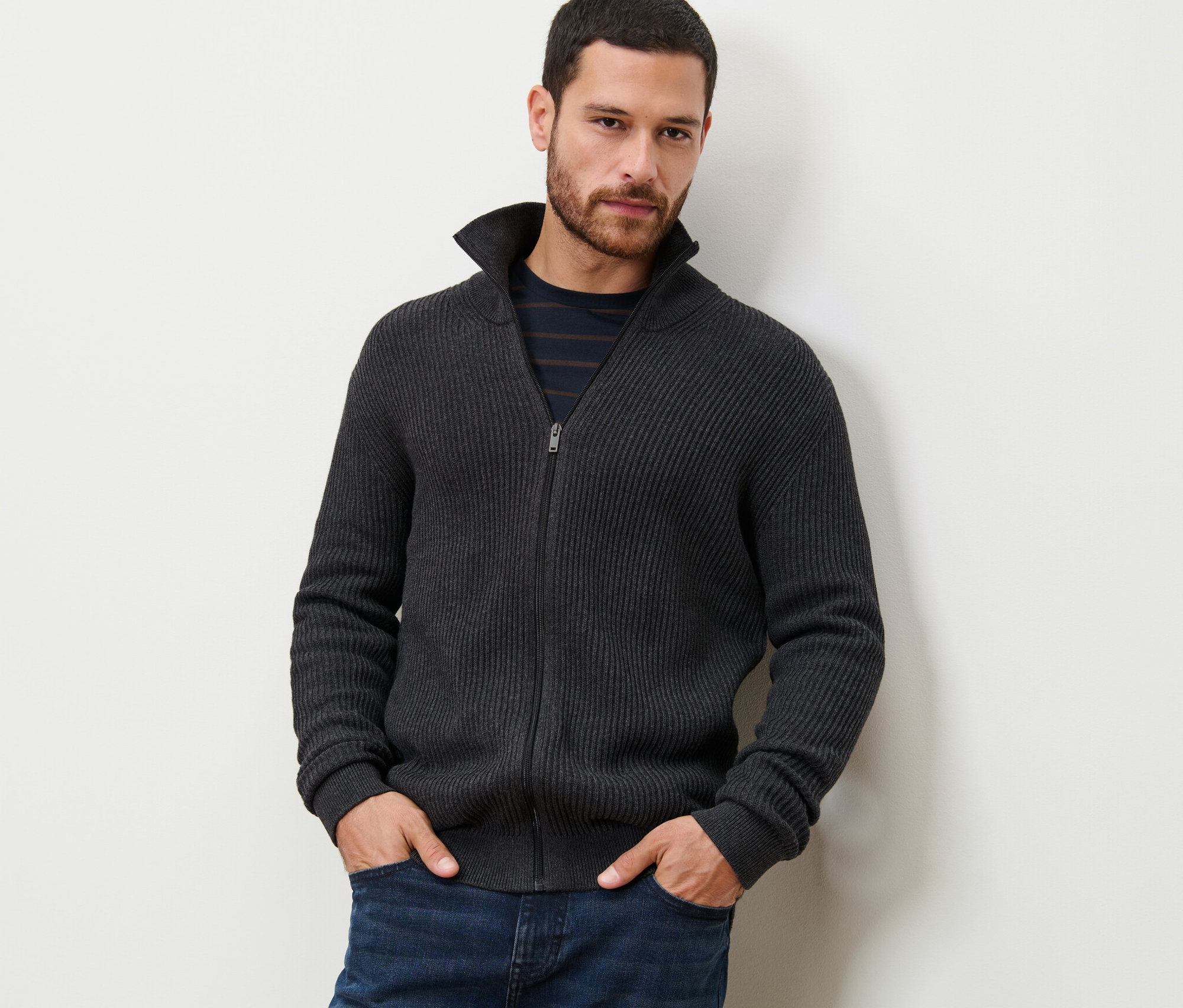 Muž stojí a má ruce v kapsách. Oblečený má pletený svetr antracitové barvy.