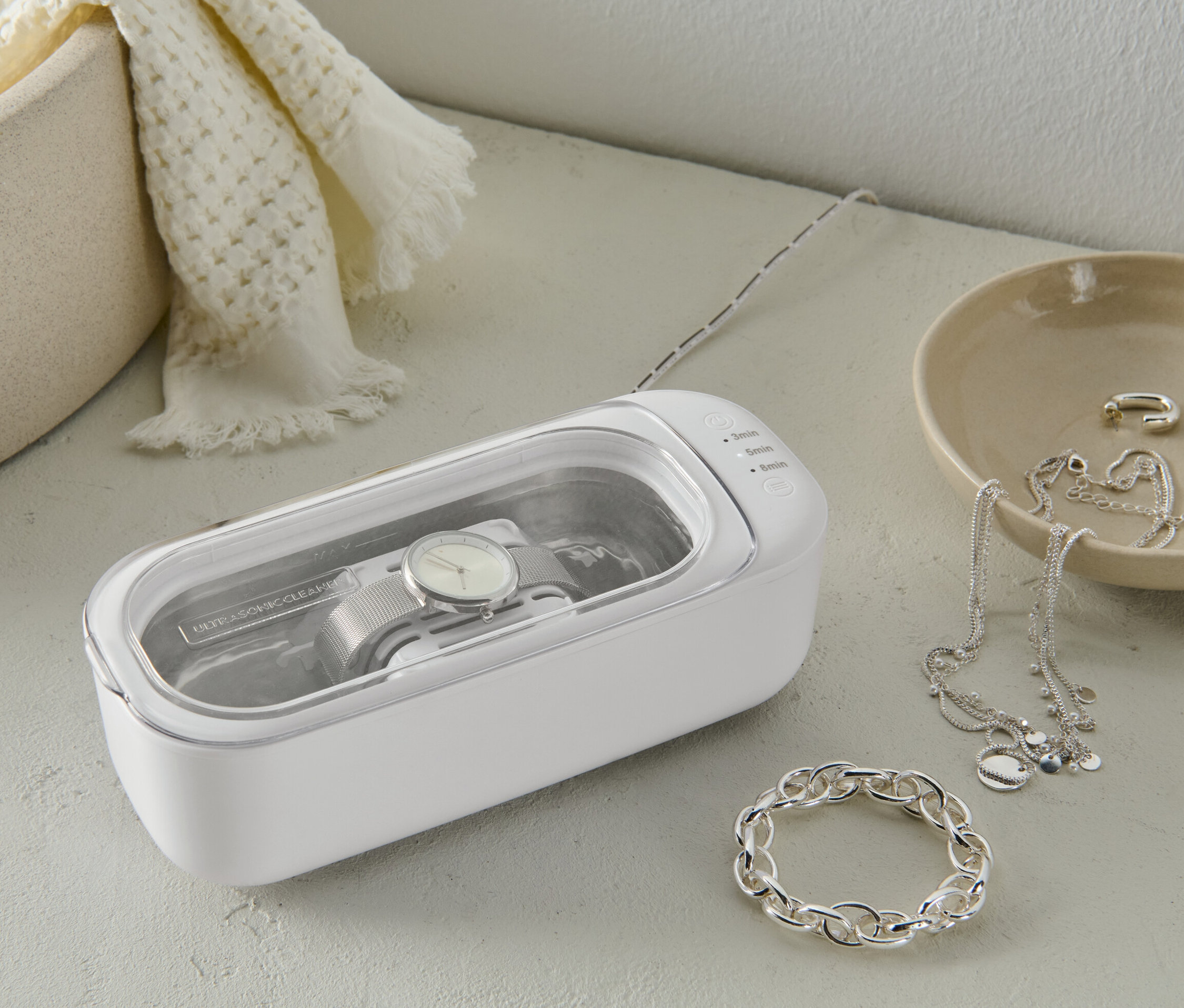 Hodinky se čistí v bílé ultrazvukové čističce. Vedle jsou šperky.