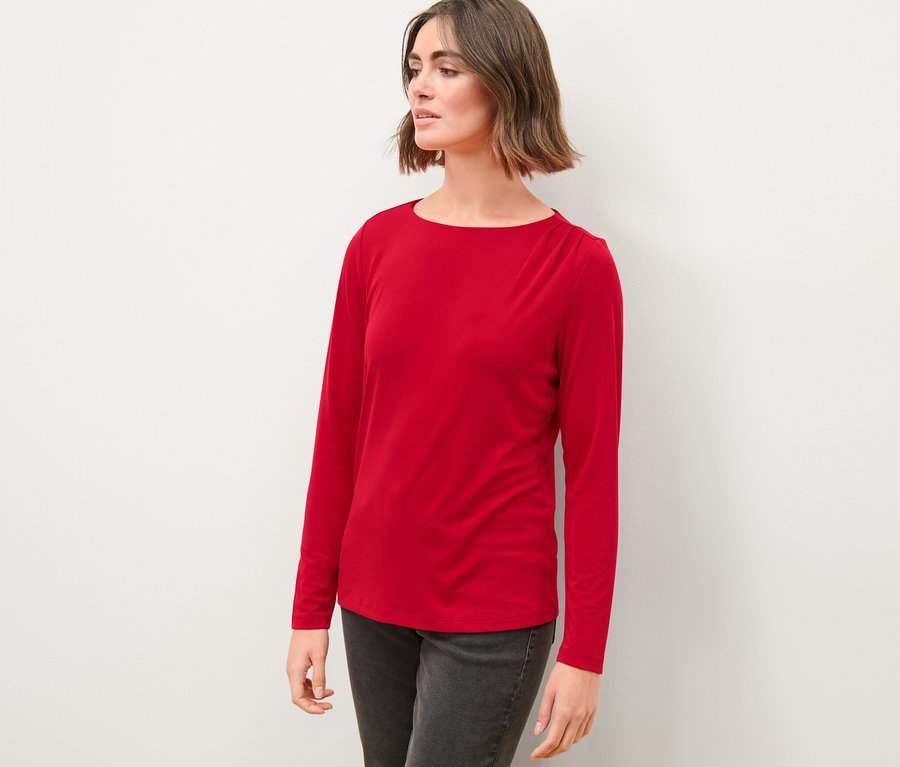 Žena v červeném halenkovém triku s řasením a šedých džínách stojí před bílou zdí.