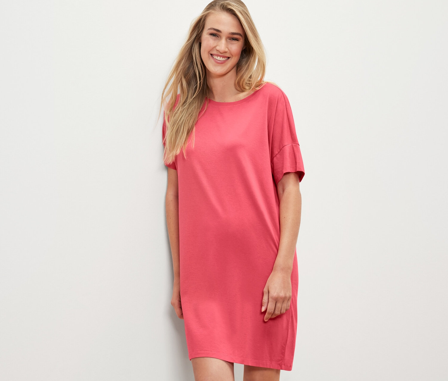 Blondýna v korálové noční košili stojí opřená o bílou zeď.