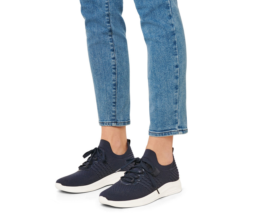 Nohy osoby v džínách a modrých teniskách slip-on s bílou podrážkou.
