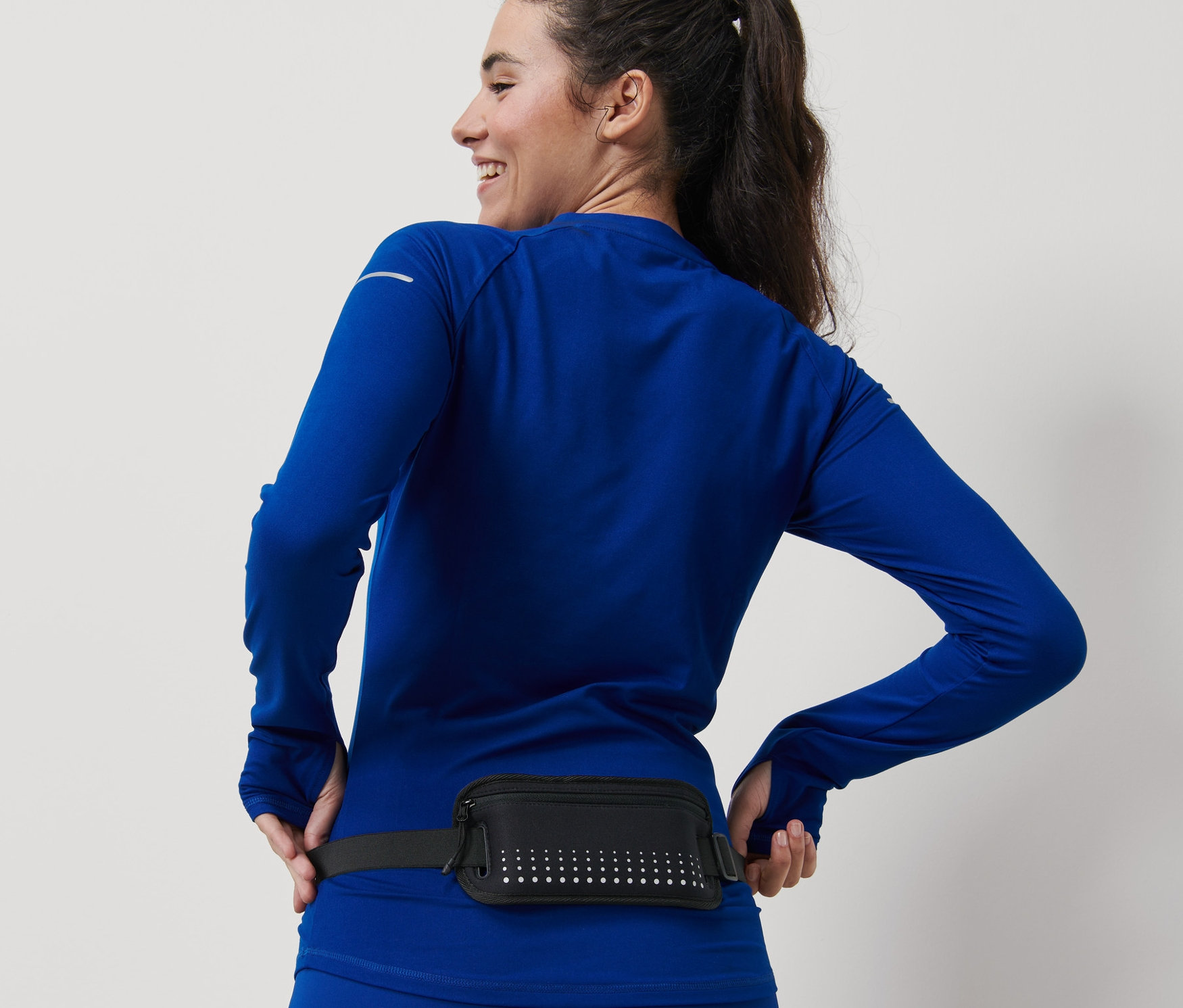Usmívající se žena v modrém sportovním oblečení má na sobě černý běžecký pás s pouzdrem na smartphone.