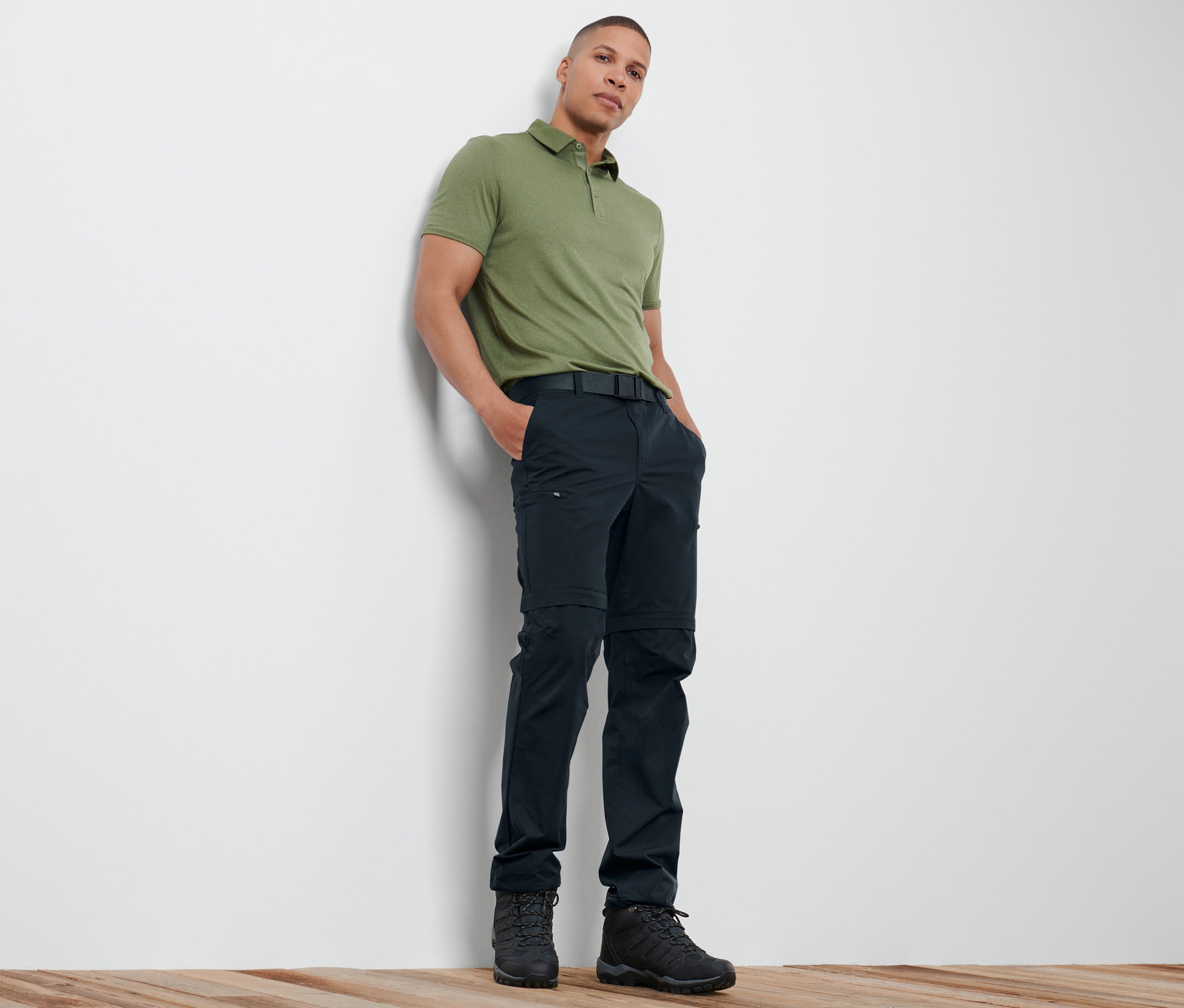 Muž opřený o stěnu má na sobě zelené polo tričko a námořnické modré funkční kalhoty 2 v 1.