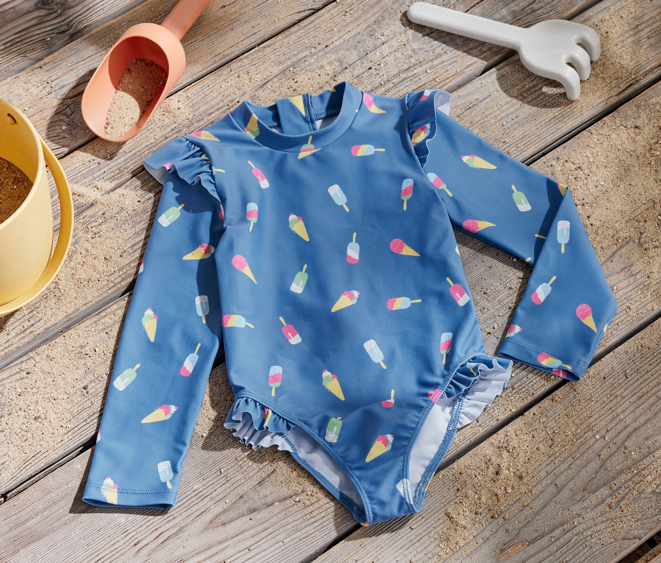 Modré dětské plavky s potiskem zmrzliny a UV faktorem 80 leží na dřevěném povrchu vedle hračky na písek.
