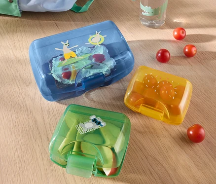 Na stole leží tři dózy na svačinu, vedle nichž je lahev s vodou a batoh.