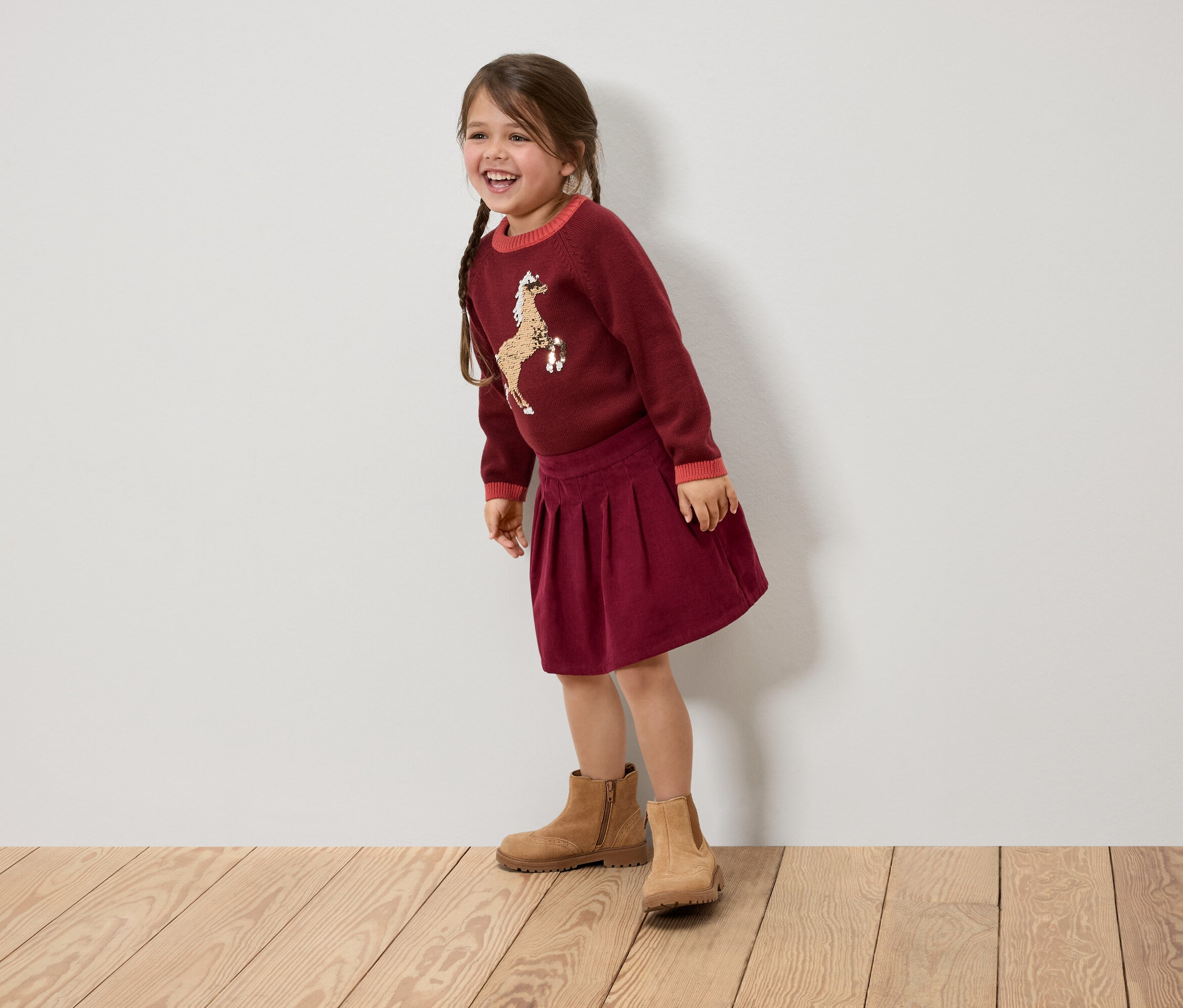 Usmívající se holčička s copánky má na sobě bordó svetr s koněm, sukni a hnědé boty.