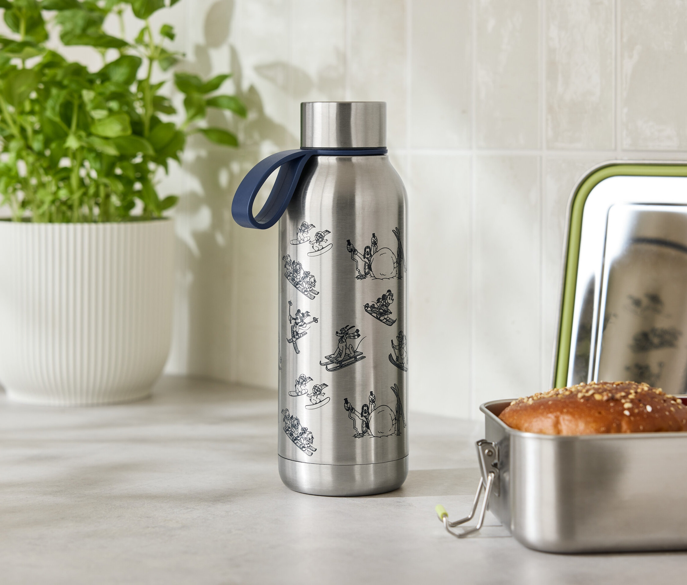 Nerezová láhev na vodu s modrým popruhem stojí vedle otevřené nerezové svačinové krabičky s houskou a rajčetem.