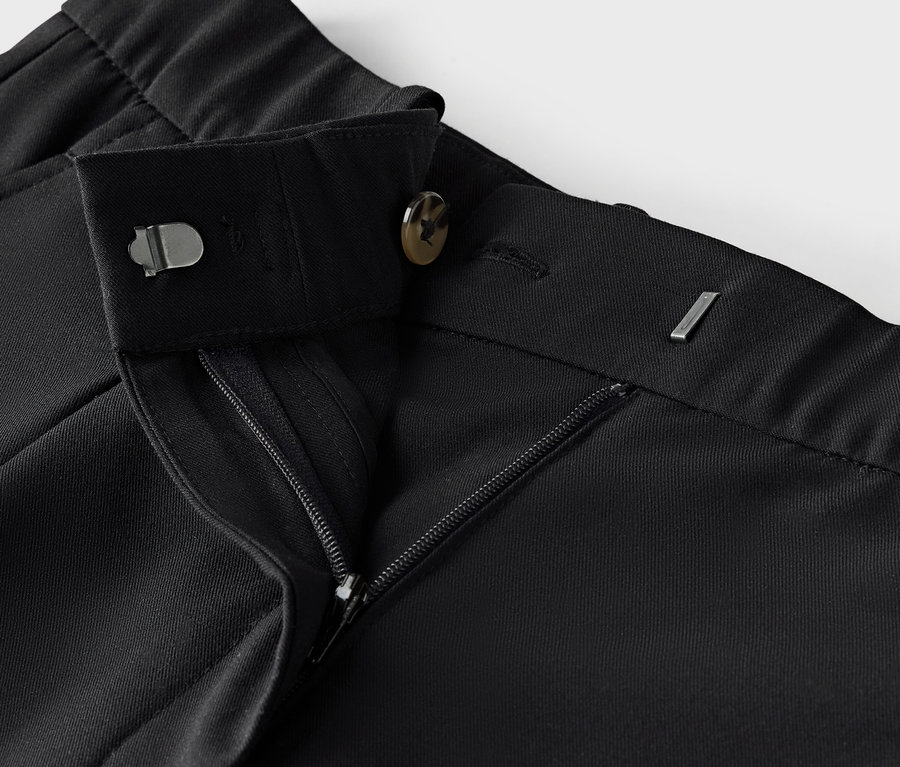 Detail černé látkové kalhoty se zipem a knoflíkem.