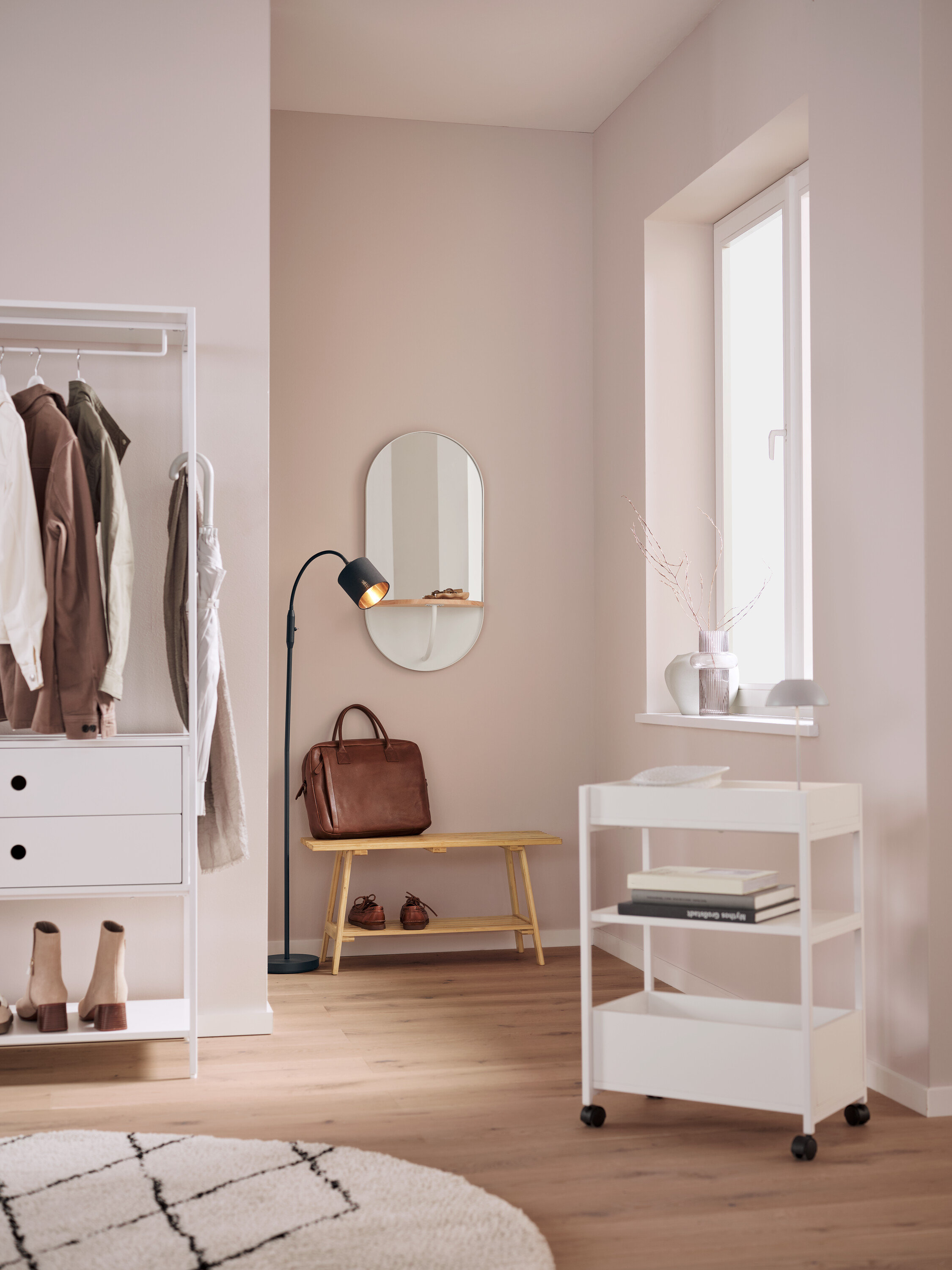 V interiéru je bílý regál s oblečením, oválné zrcadlo, hnědá kožená taška na lavici, černá lampa a bílý pojízdný vozík.