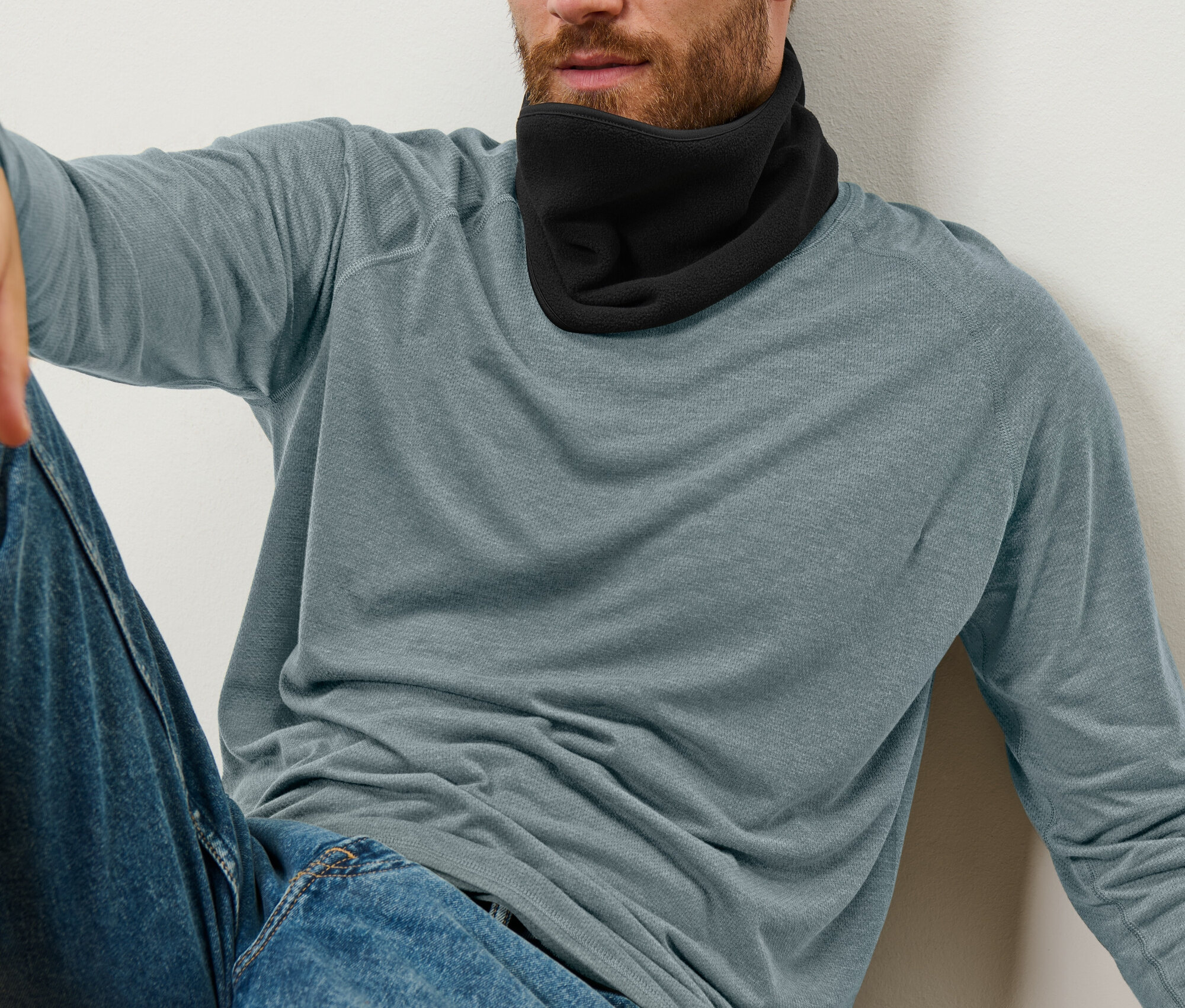 Muž sedí opřený o stěnu a má na sobě černý nákrčník, modré džíny a modrošedé tričko s dlouhým rukávem.