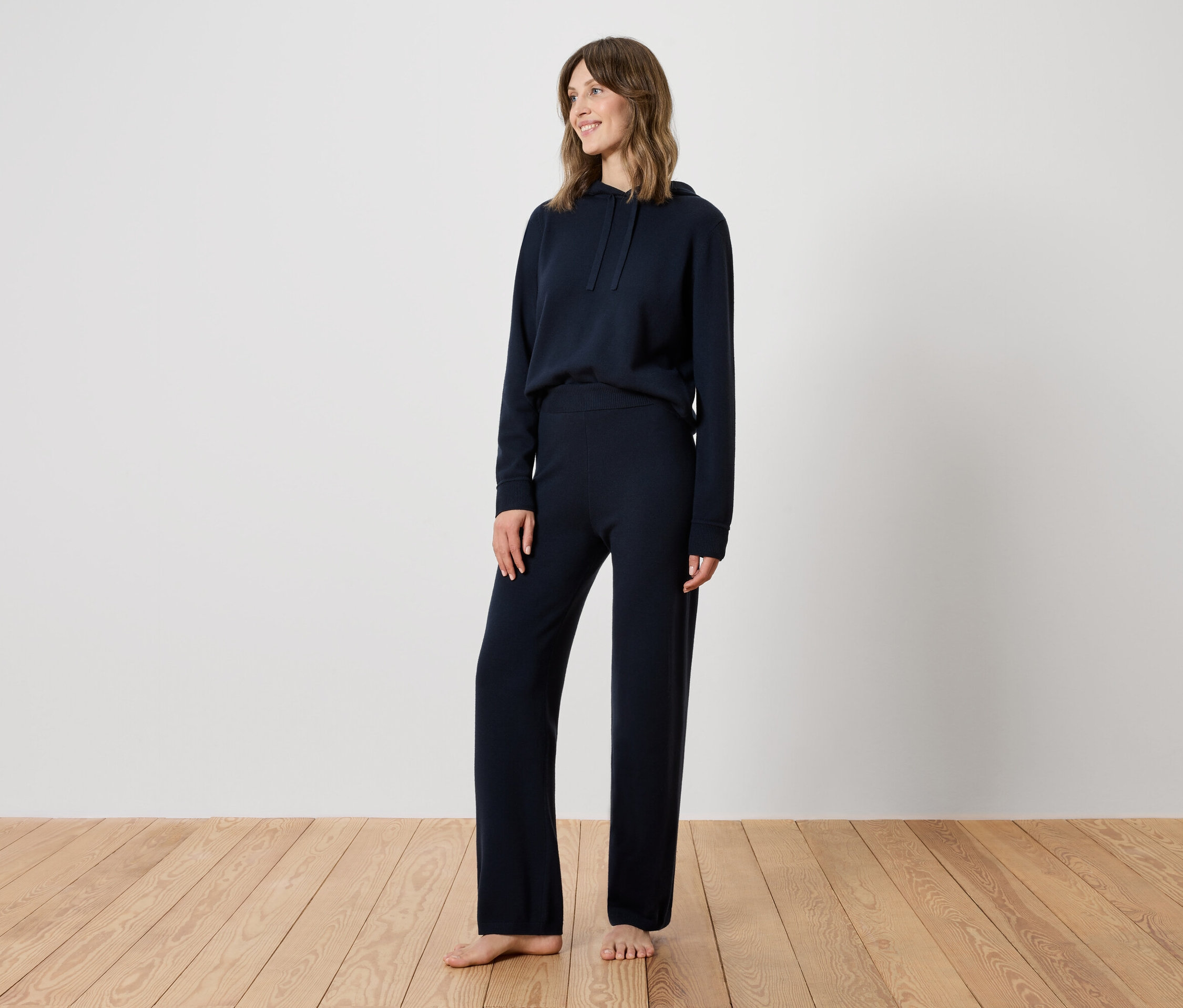 Žena stojí na dřevěné podlaze a má na sobě tmavě modrou teplákovou soupravu.