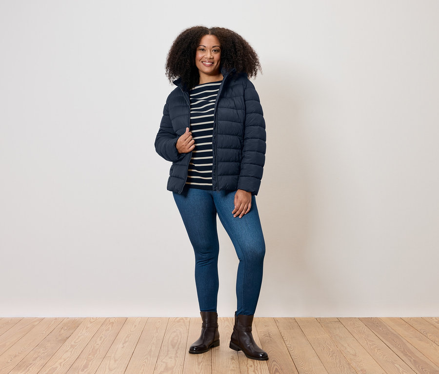 Žena s kudrnatými vlasy stojí a má na sobě modrou prošívanou bundu, pruhované tričko, džíny a boty.