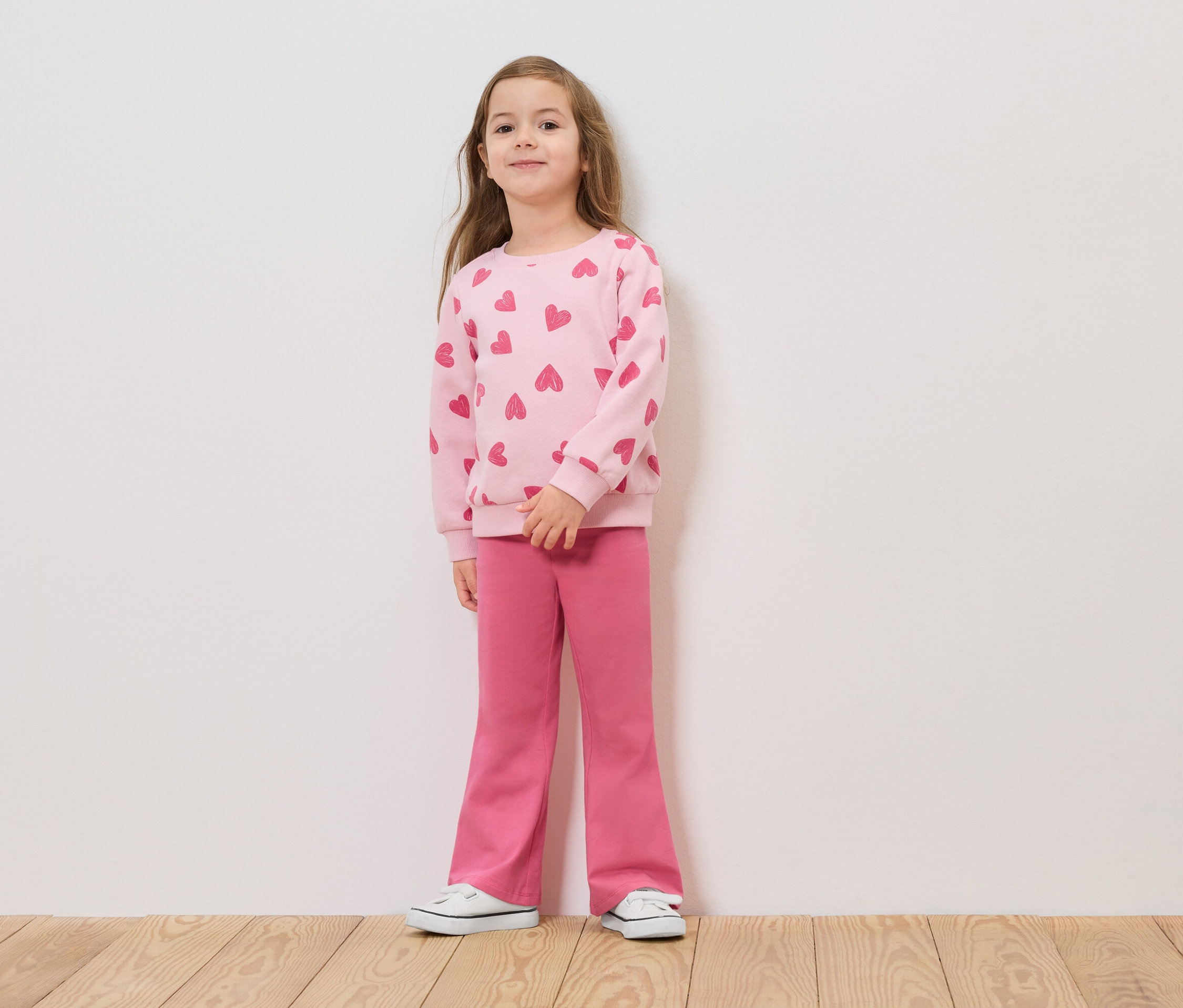 Dívka stojí před bílou zdí v růžovém svetru se srdíčky a růžových kalhotách.