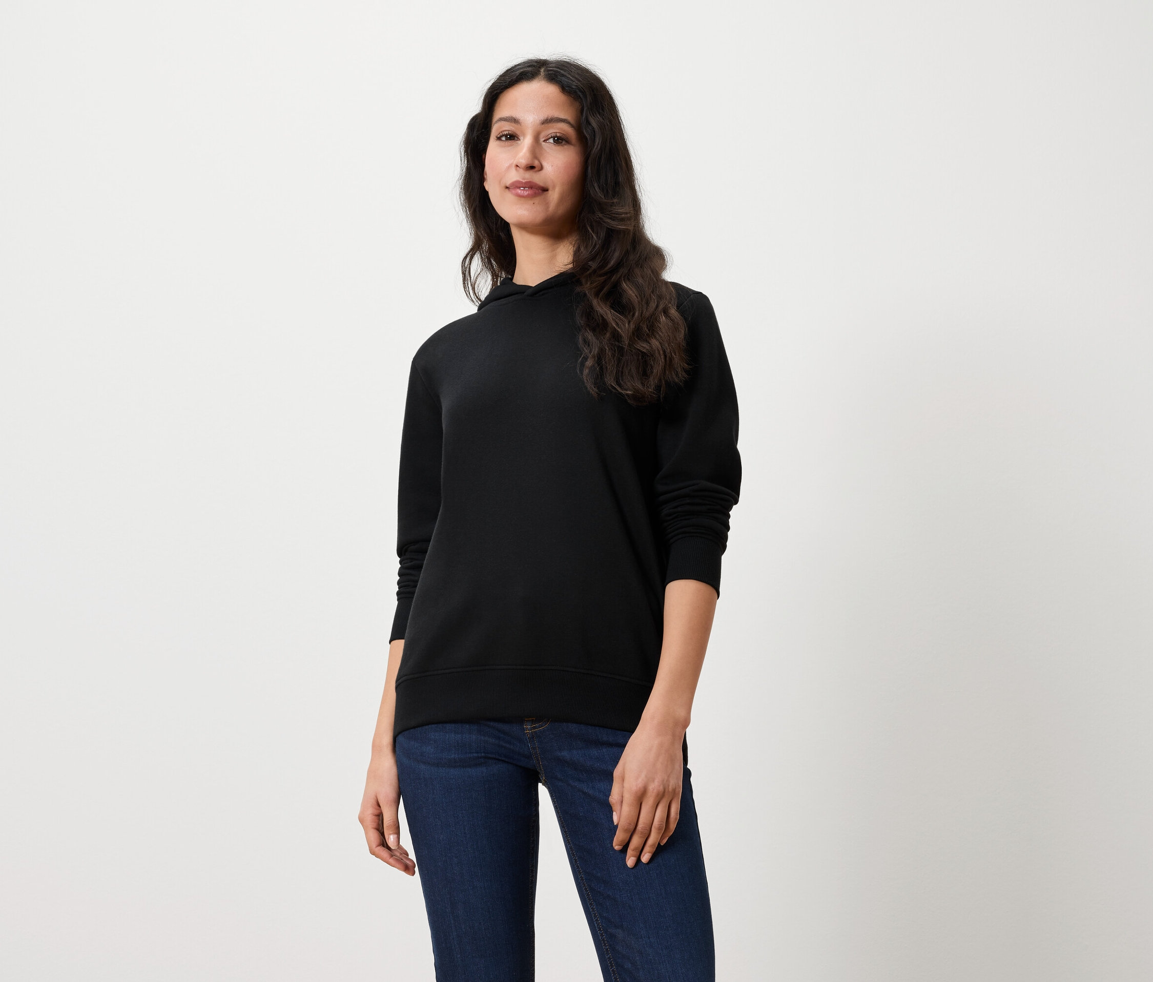 Žena s dlouhými hnědými vlasy stojí a má na sobě černou mikinu s kapucí a džíny.