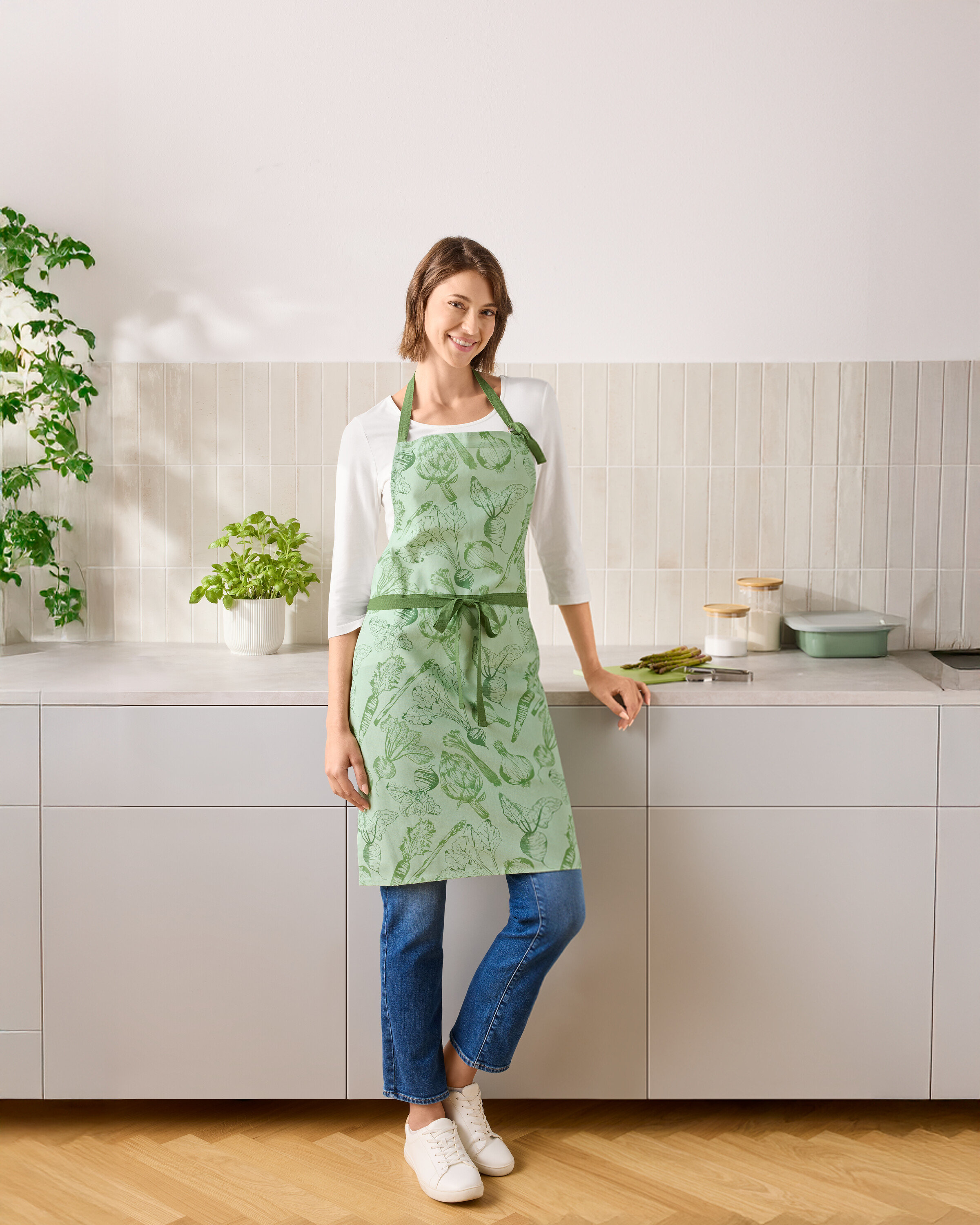 Žena stojí v kuchyni a má na sobě zelenou zástěru se zeleninou.