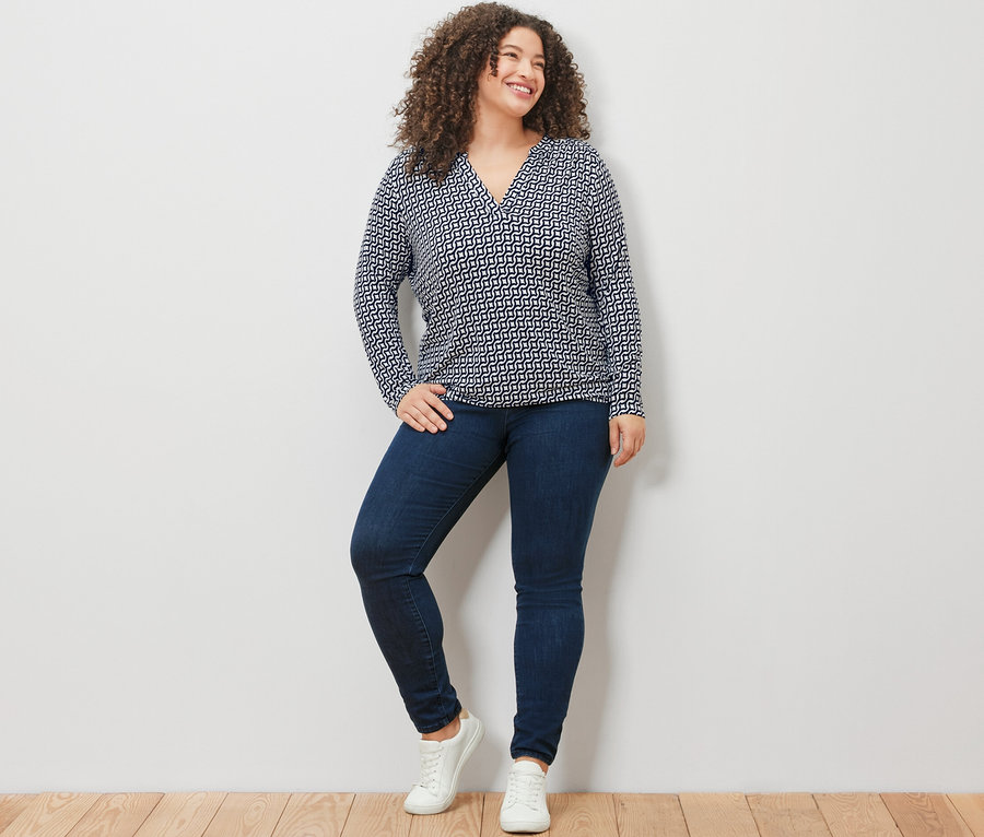 Žena stojící před bílou zdí má na sobě námořnicky modré halenkové triko s potiskem, džíny a bílé tenisky.