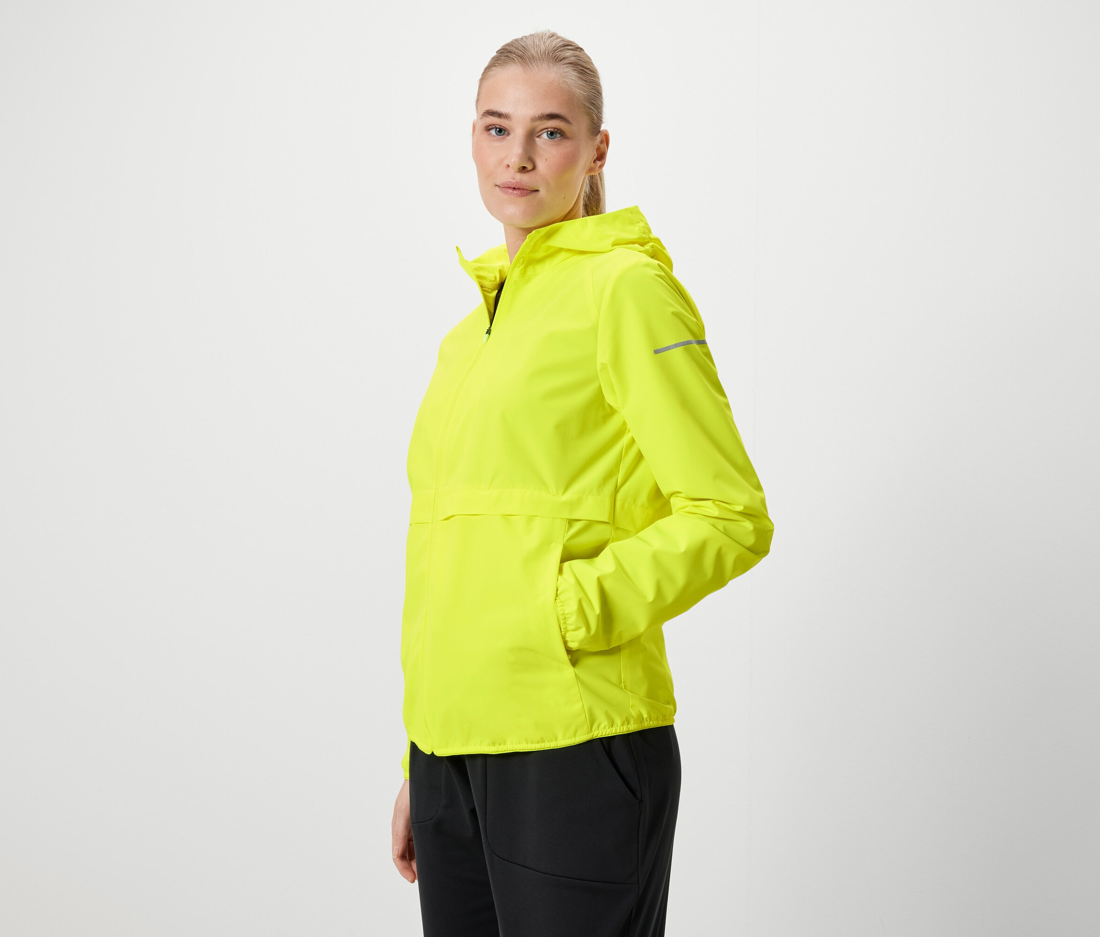 Blondýna v neonově žluté bundě s kapucí a černých kalhotách pózuje bokem.