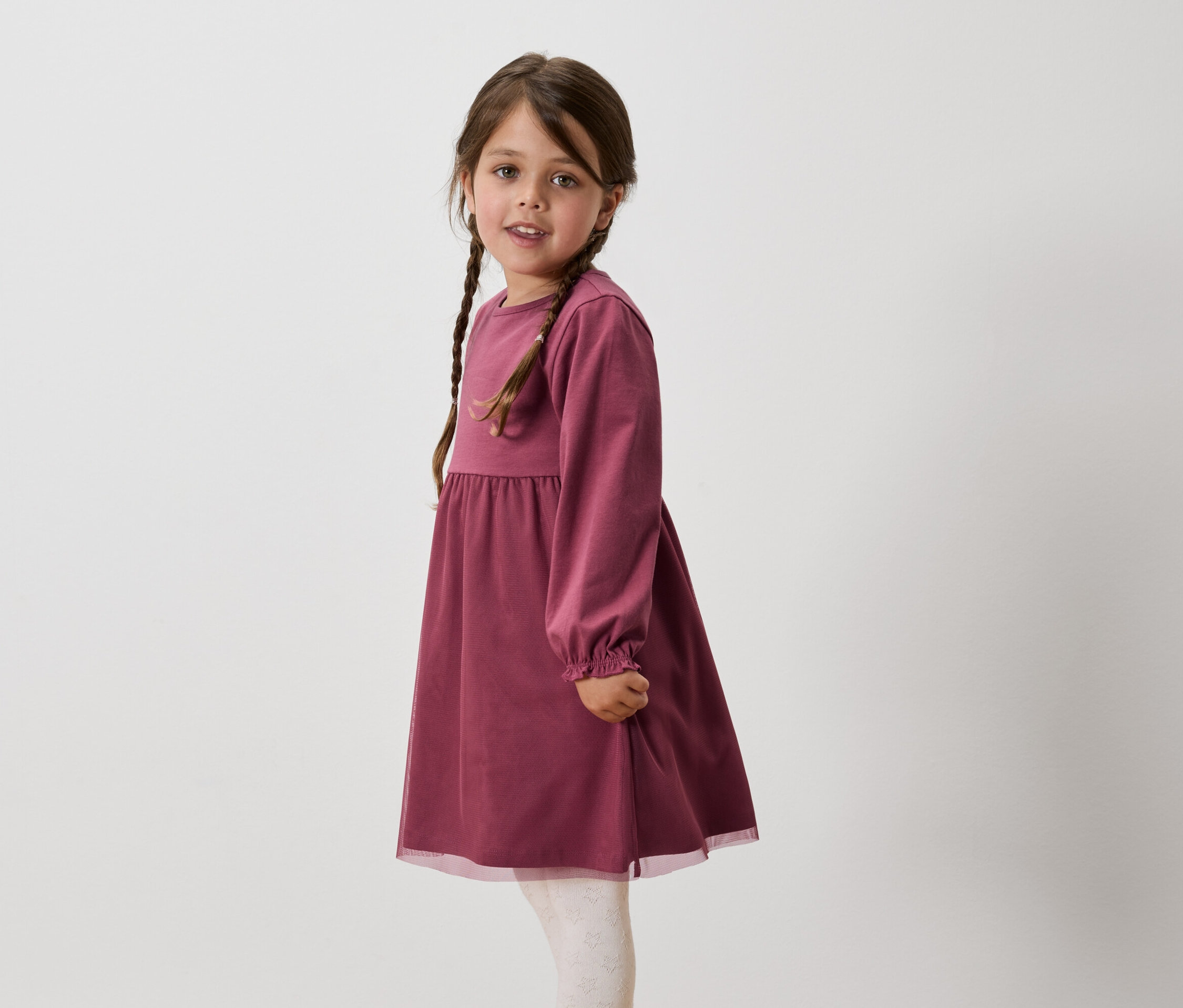 Dívka s hnědými vlasy spletenými do copů má na sobě vínové šaty s dlouhým rukávem a bílé legíny.