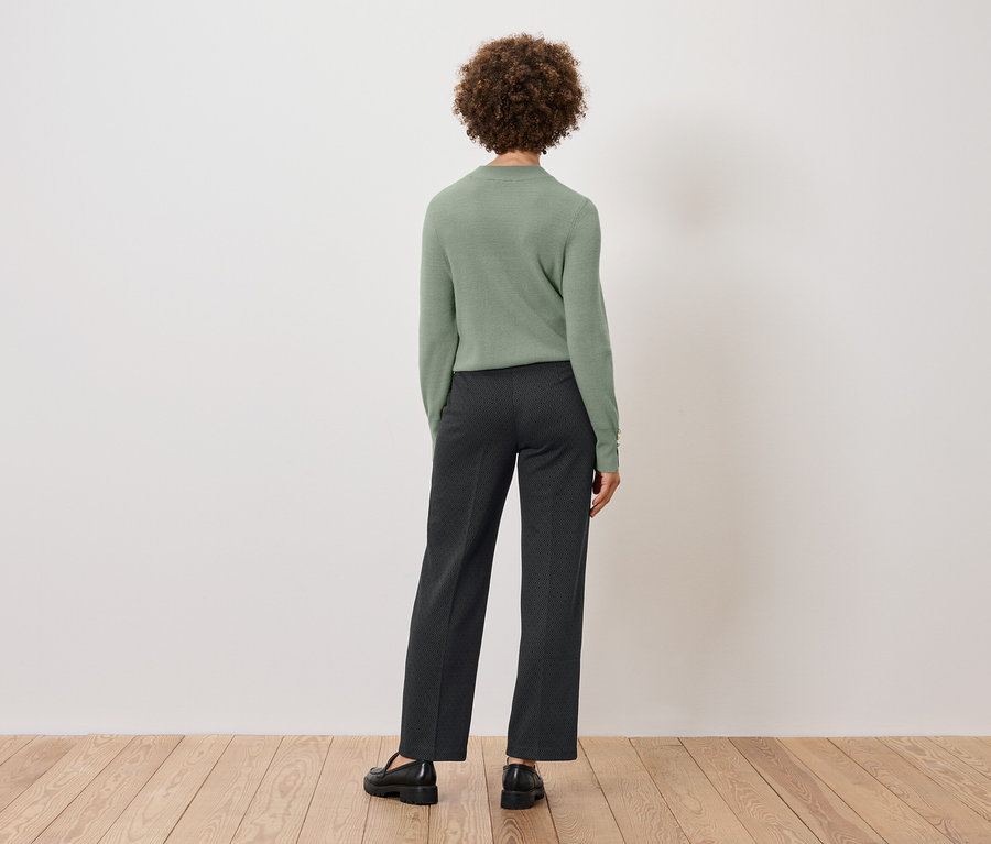 Zezadu vidíme ženu s kudrnatými vlasy v zeleném svetru a tmavých kalhotách, stojící na dřevěné podlaze před bílou zdí.