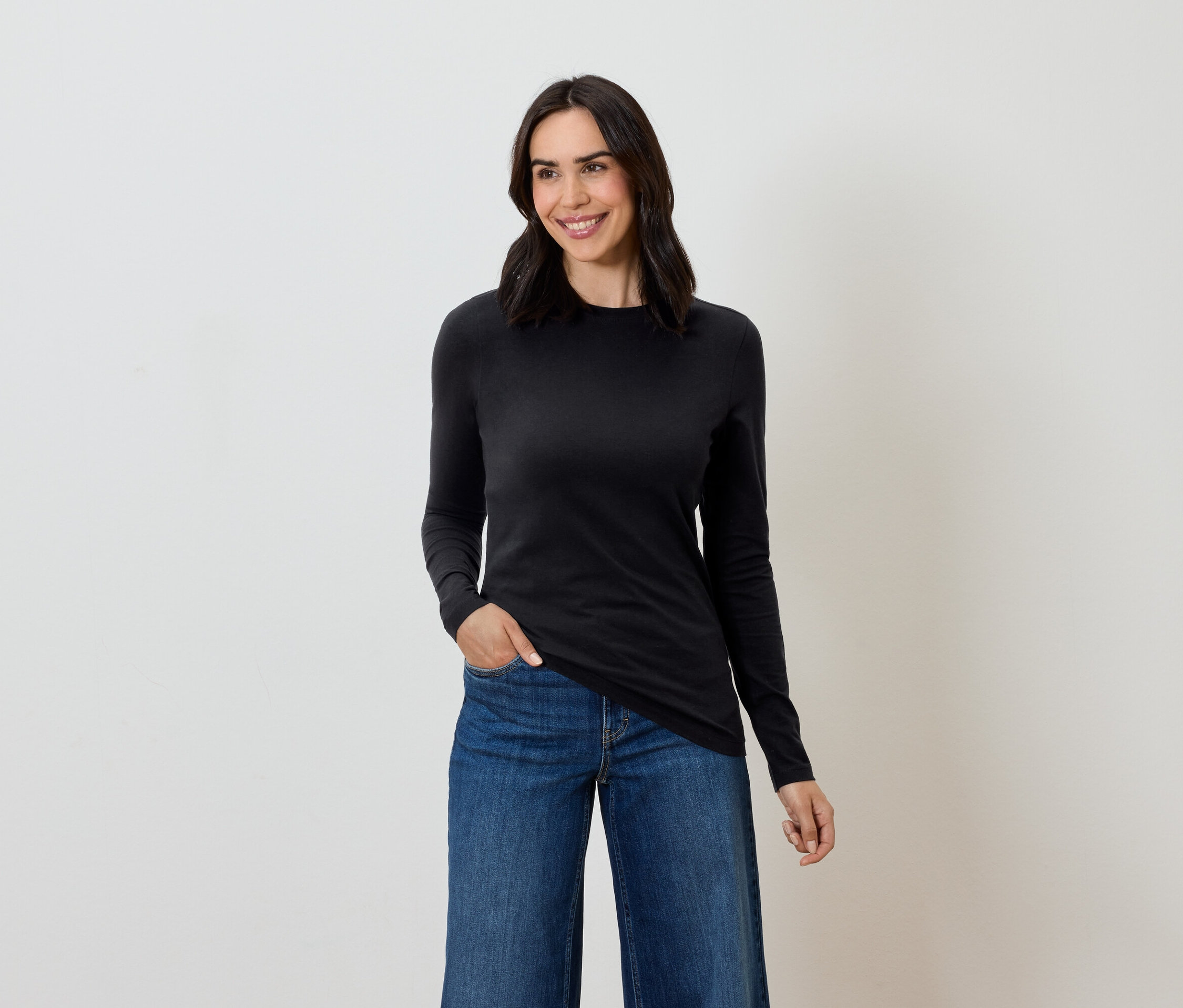 Žena s hnědými vlasy pózuje v černém tričku s dlouhým rukávem a modrých džínách.