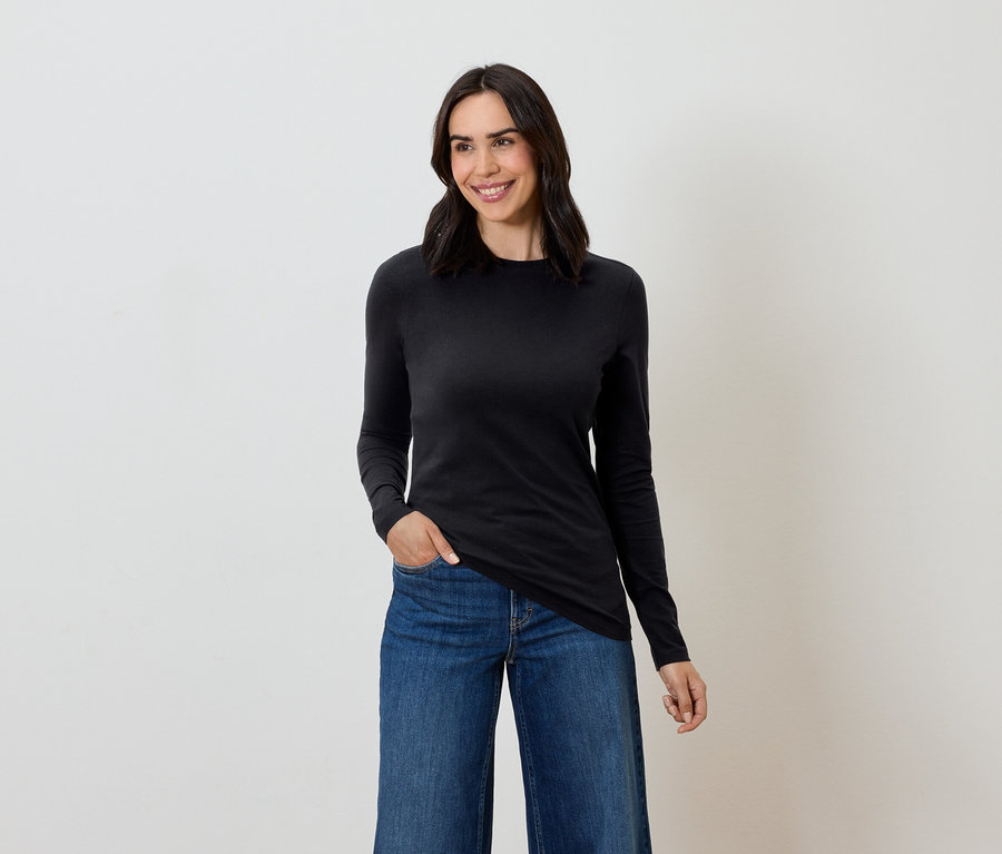 Žena s hnědými vlasy pózuje v černém tričku s dlouhým rukávem a modrých džínách.