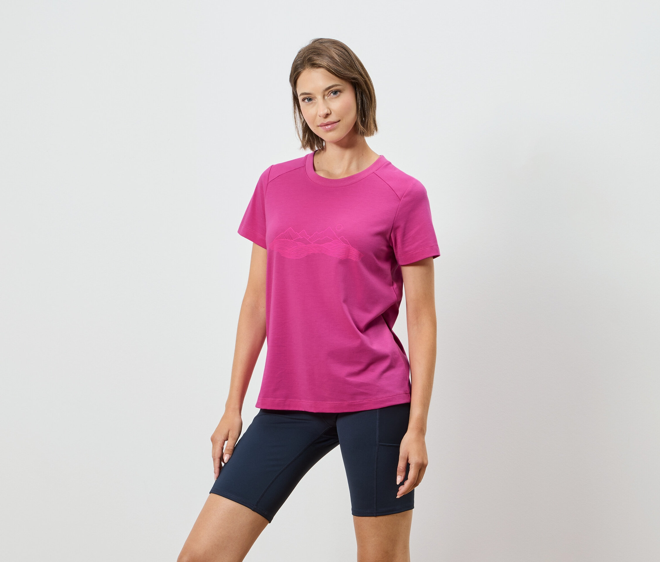 Žena má na sobě růžové tričko s horským motivem a tmavě modré šortky na bílém pozadí.