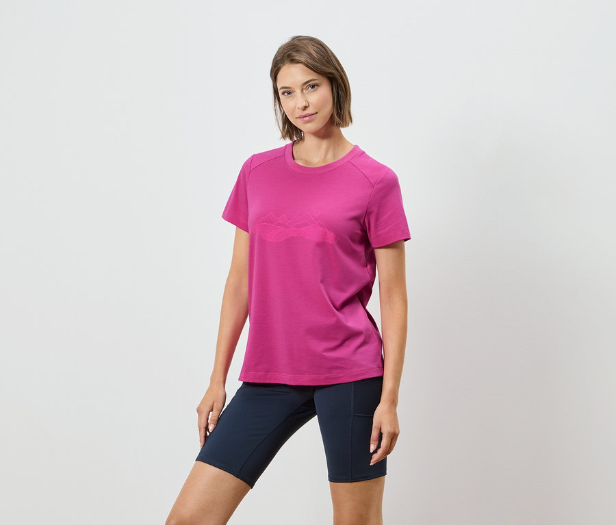 Žena má na sobě růžové tričko s horským motivem a tmavě modré šortky na bílém pozadí.
