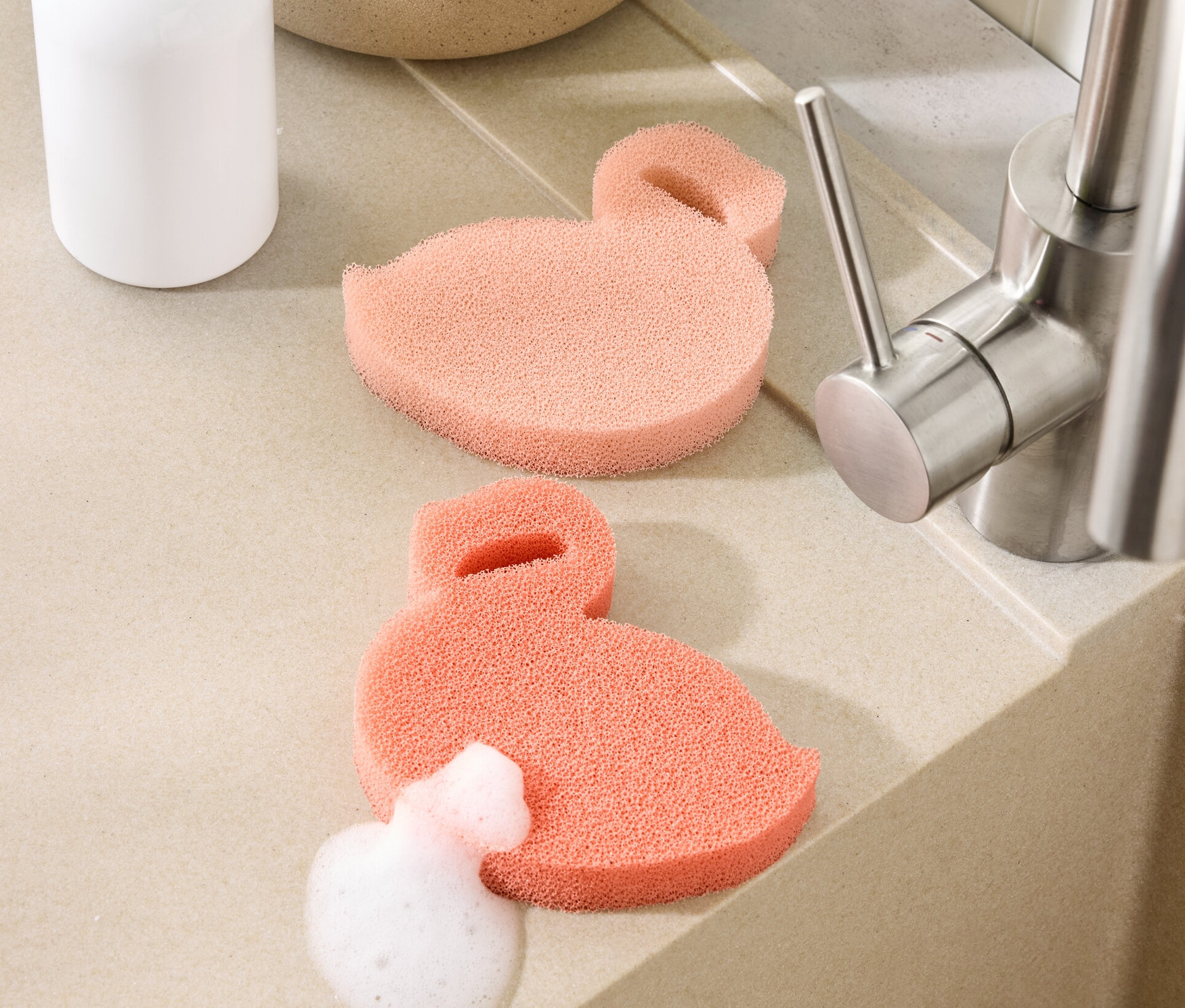 Dvě silikonové houbičky ve tvaru plameňáka leží na lince u dřezu. Jedna z nich je pokrytá pěnou.