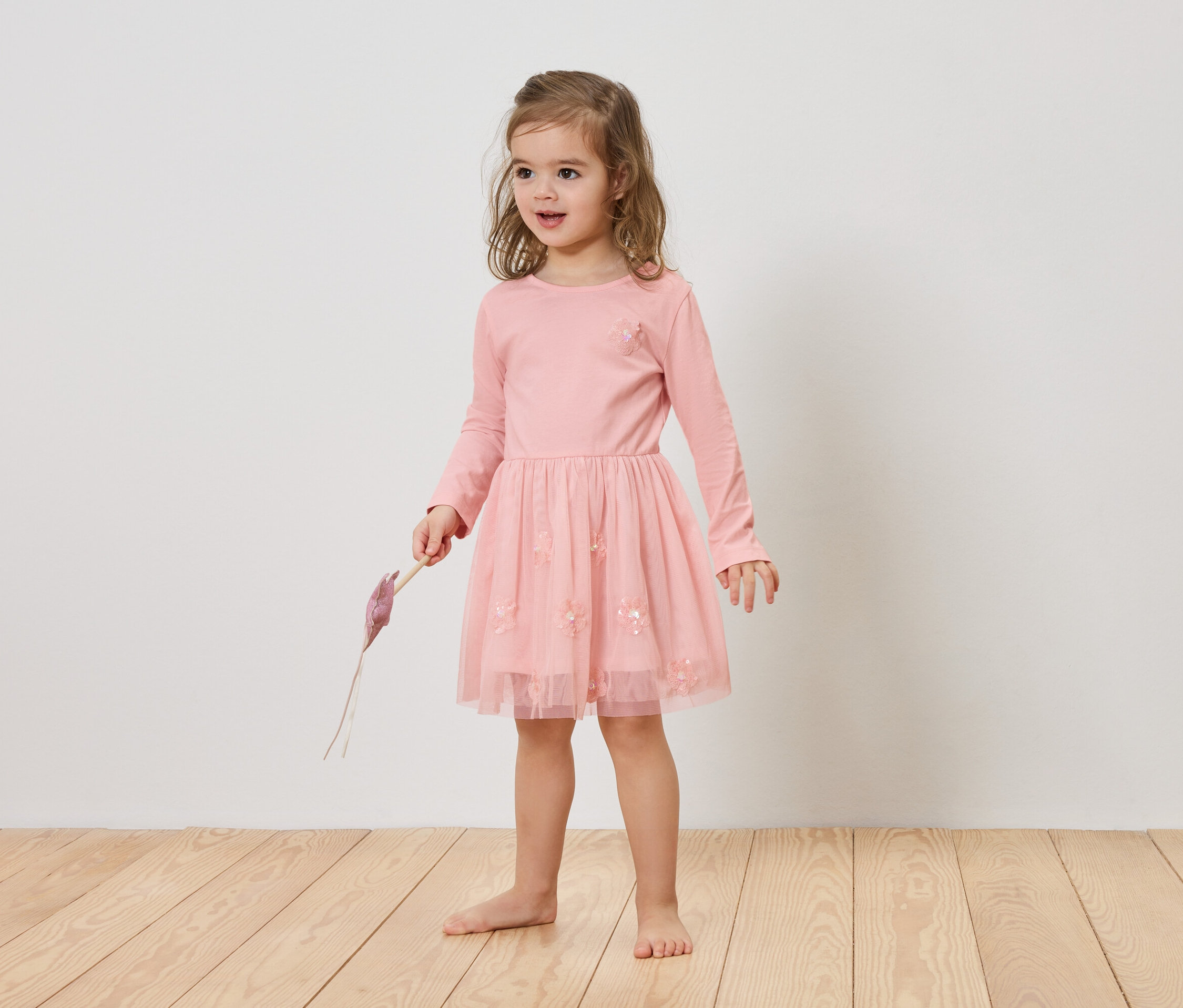 Dívka s blond vlasy má na sobě růžové šaty a drží v ruce fialový předmět.