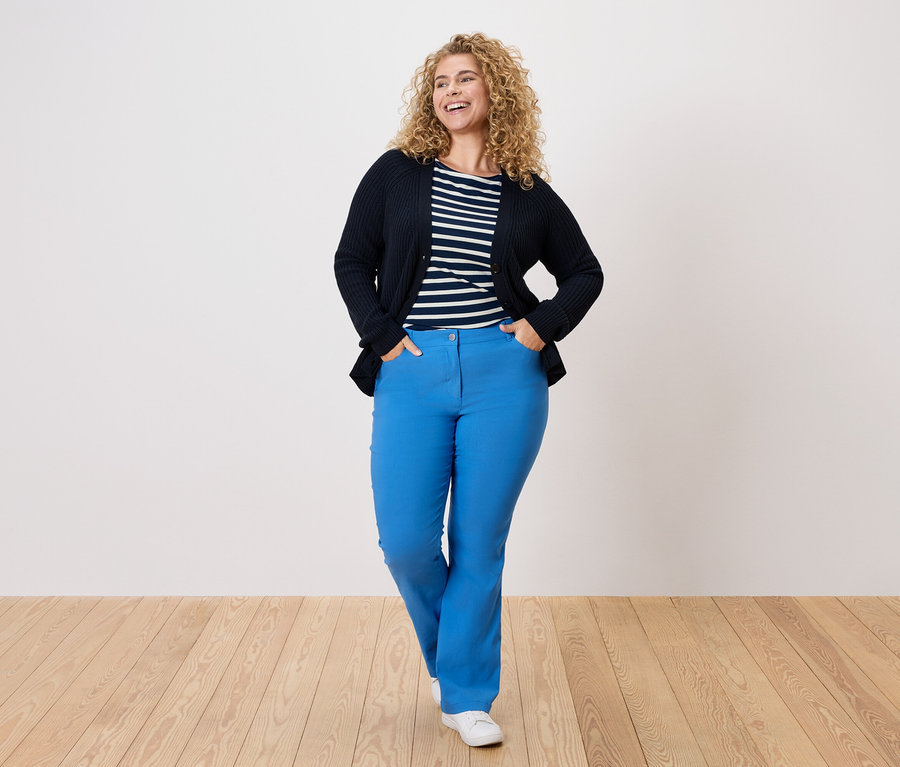 Žena s kudrnatými vlasy má na sobě tmavě modrý kardigan, pruhovanou košili a modré kalhoty.