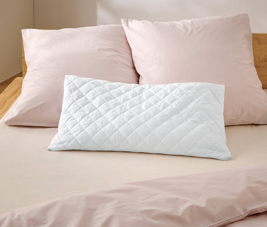 Na posteli leží dva růžové polštáře a jeden bílý prošívaný polštář.