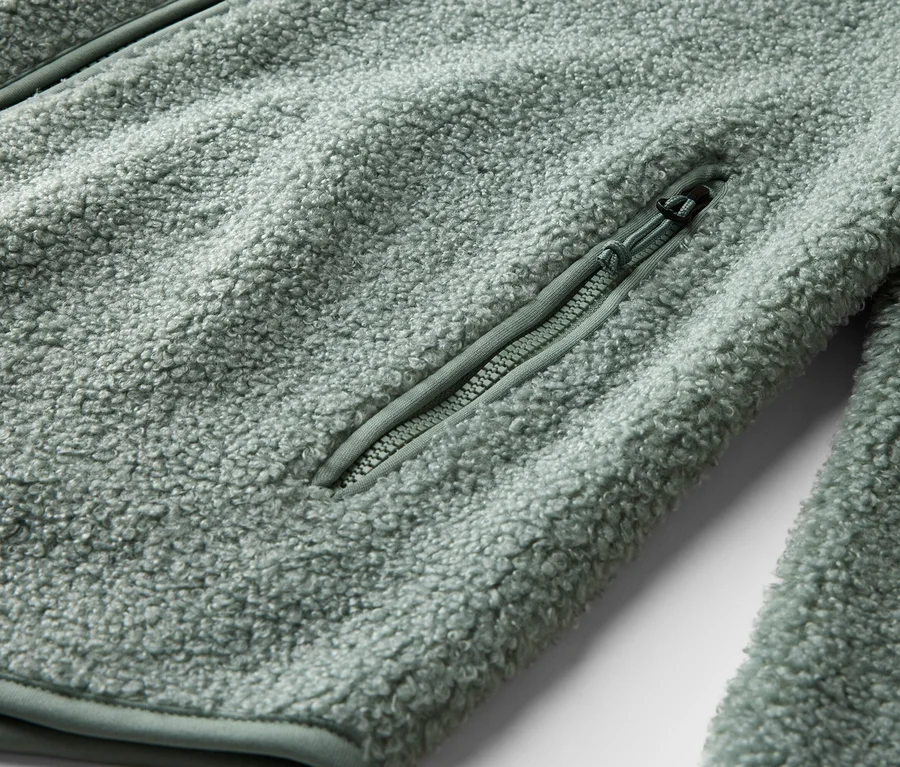 Detailní záběr světle zelené fleecové bundy s kapsou na zip.